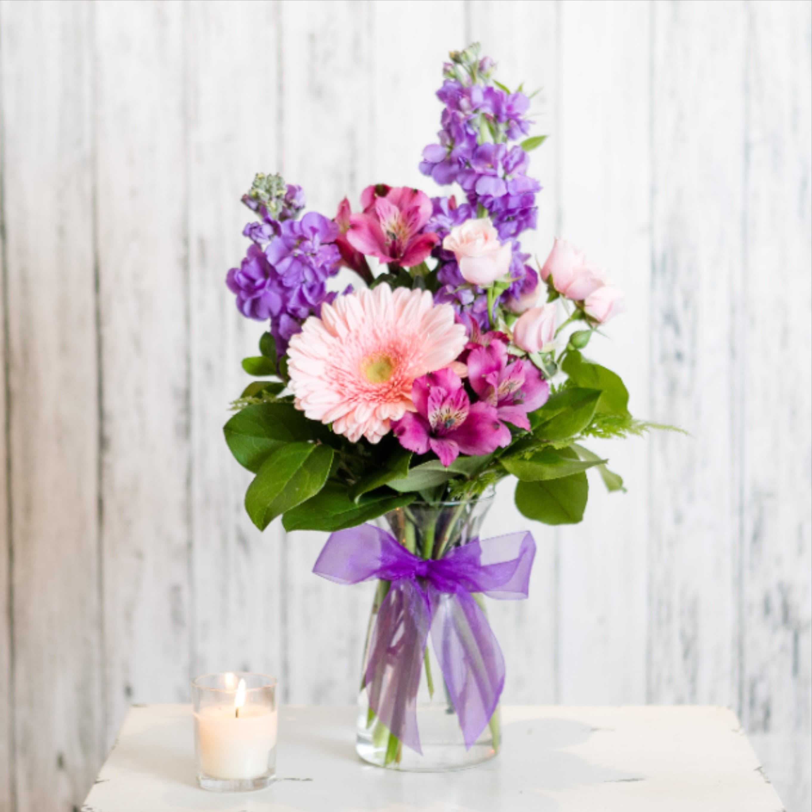 Mixed bouquet of pink and purple flowers in a clear vase with purple bow beside a lit candle.