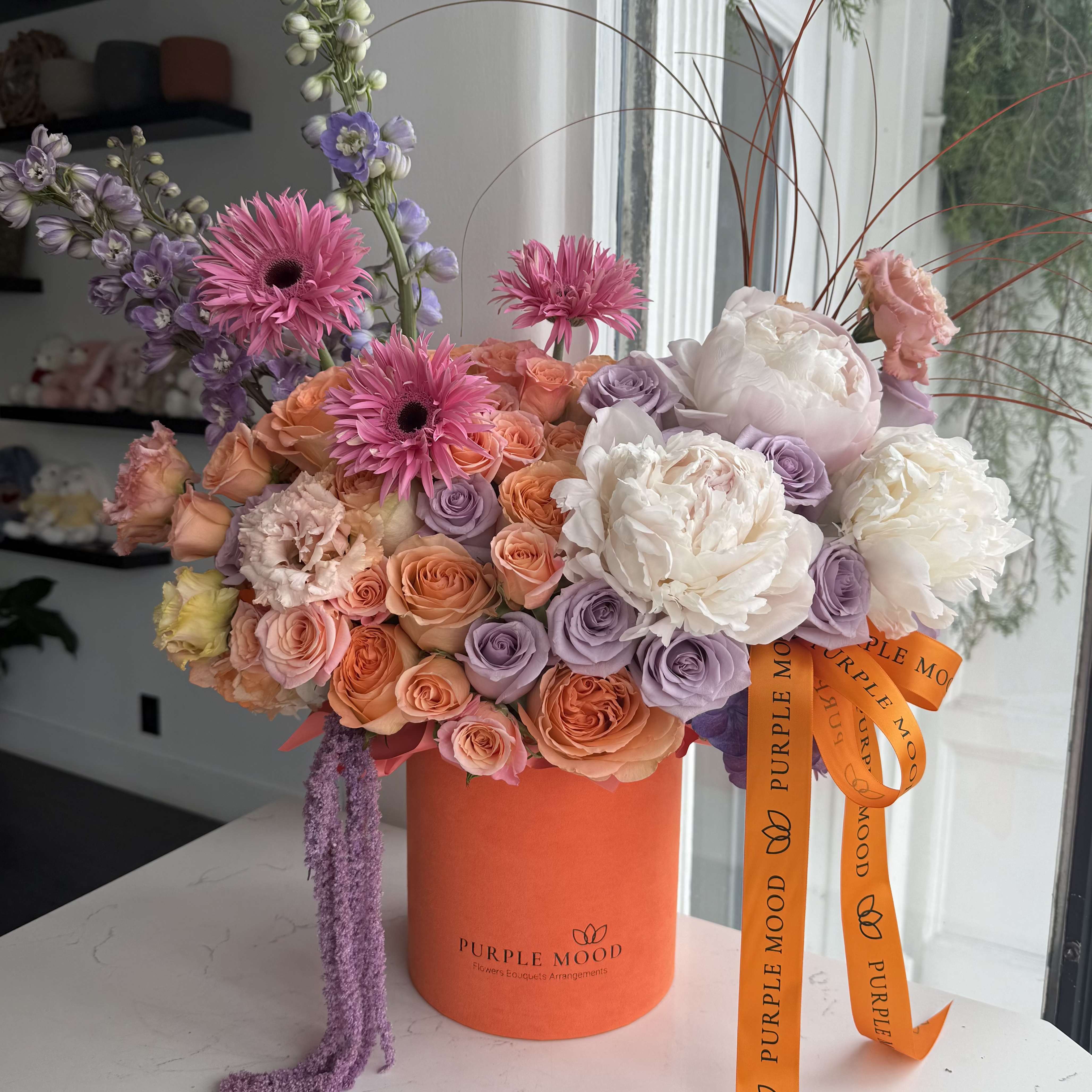 Aperol Bloom Box - A vibrant and refreshing floral arrangement inspired by the iconic Aperol spritz palette. Designed in warm peach, coral, and soft lavender tones, this box features premium garden roses, spray roses, airy delphinium, and seasonal blooms for a lively, sun-kissed look.  The box is designed with a back lid and a fresh green interior, creating a beautiful contrast that makes the blooms stand out even more.  Perfect for celebrations, gifting, or adding a pop of color to any space.