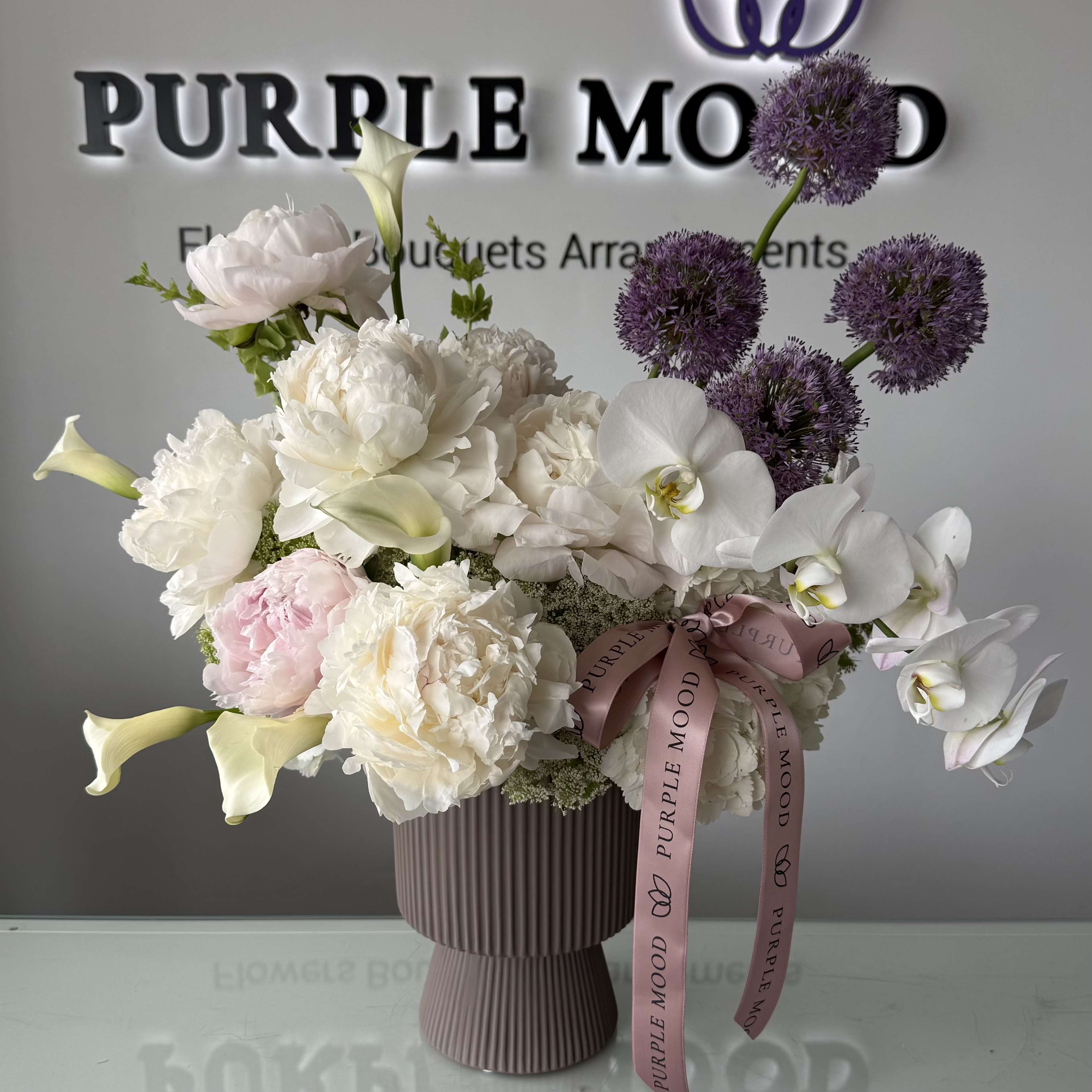 Peony Orchid Vase - A refined and elegant arrangement featuring lush peonies, delicate orchids, and premium seasonal blooms in soft ivory and blush tones. Designed with airy texture and graceful movement, this piece brings a timeless, luxurious feel to any space.  Arranged in a modern ribbed vase and finished with our signature Purple Mood ribbon. Perfect for special occasions or as a sophisticated everyday statement.  Vase included.
