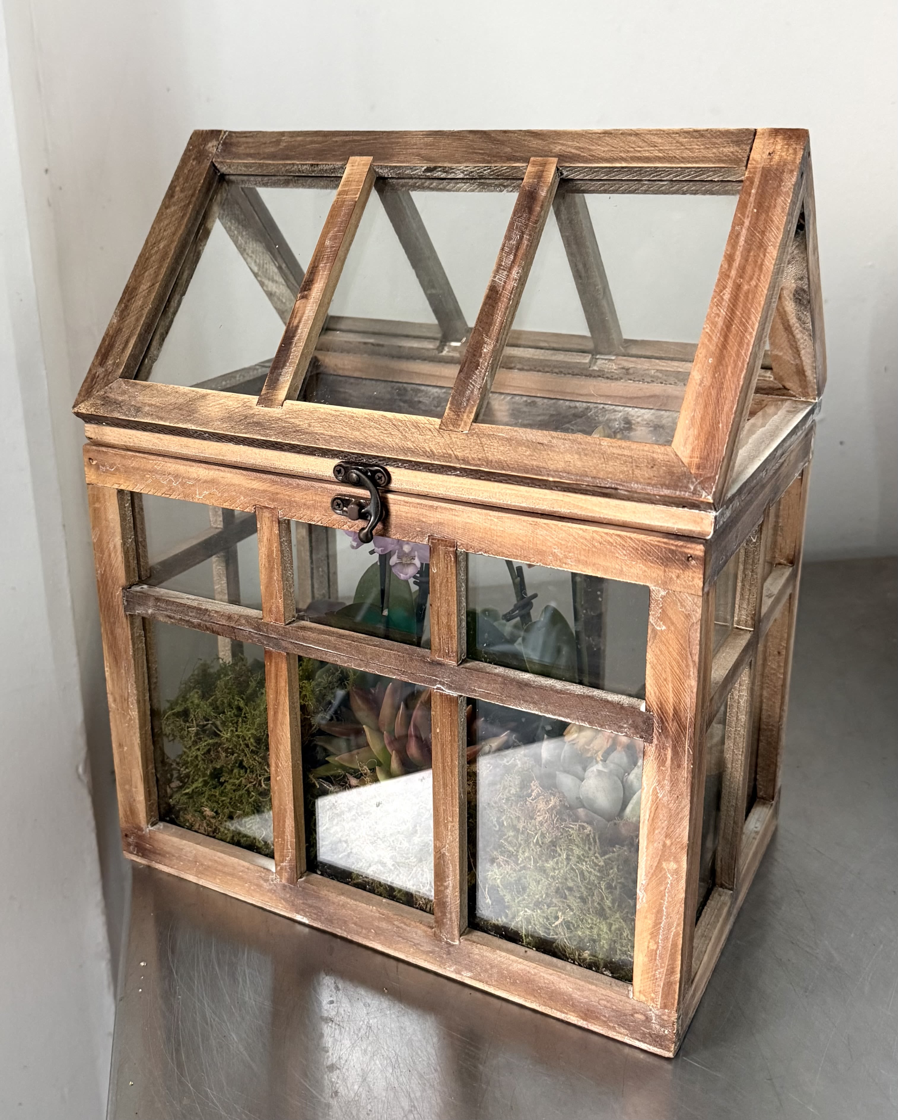Rustic Orchid &amp; Succulent Glass Greenhouse - Transform your home or office into a miniature oasis with this versatile Wardian case. Its generous height and transparent walls allow for maximum light exposure, while the secure lid helps maintain the humidity levels needed for tropical varieties like orchids and ferns. Whether as a lush indoor garden centerpiece or a vintage-inspired display box, it seamlessly combines industrial-style lines with the warmth of natural materials.  This Large Rustic Wood &amp; Glass Terrarium brings a chic farmhouse touch to any indoor space. Designed as a decorative tabletop greenhouse, it features a weathered wood frame and clear paned glass, providing a perfect environment for delicate plants like orchids and succulents.   Product Highlights:   Design: Classic house-shaped architecture with a rustic, dark-stained wood finish.  Access: Features a wide, hinged roof for easy planting, watering, and misting.  Versatility: Ideal for live or faux plants, fairy gardens, air plants, or even as a unique candle lantern.  Multi-Purpose: Frequently used beyond gardening as an elegant wedding card box or a display case for collectibles.  