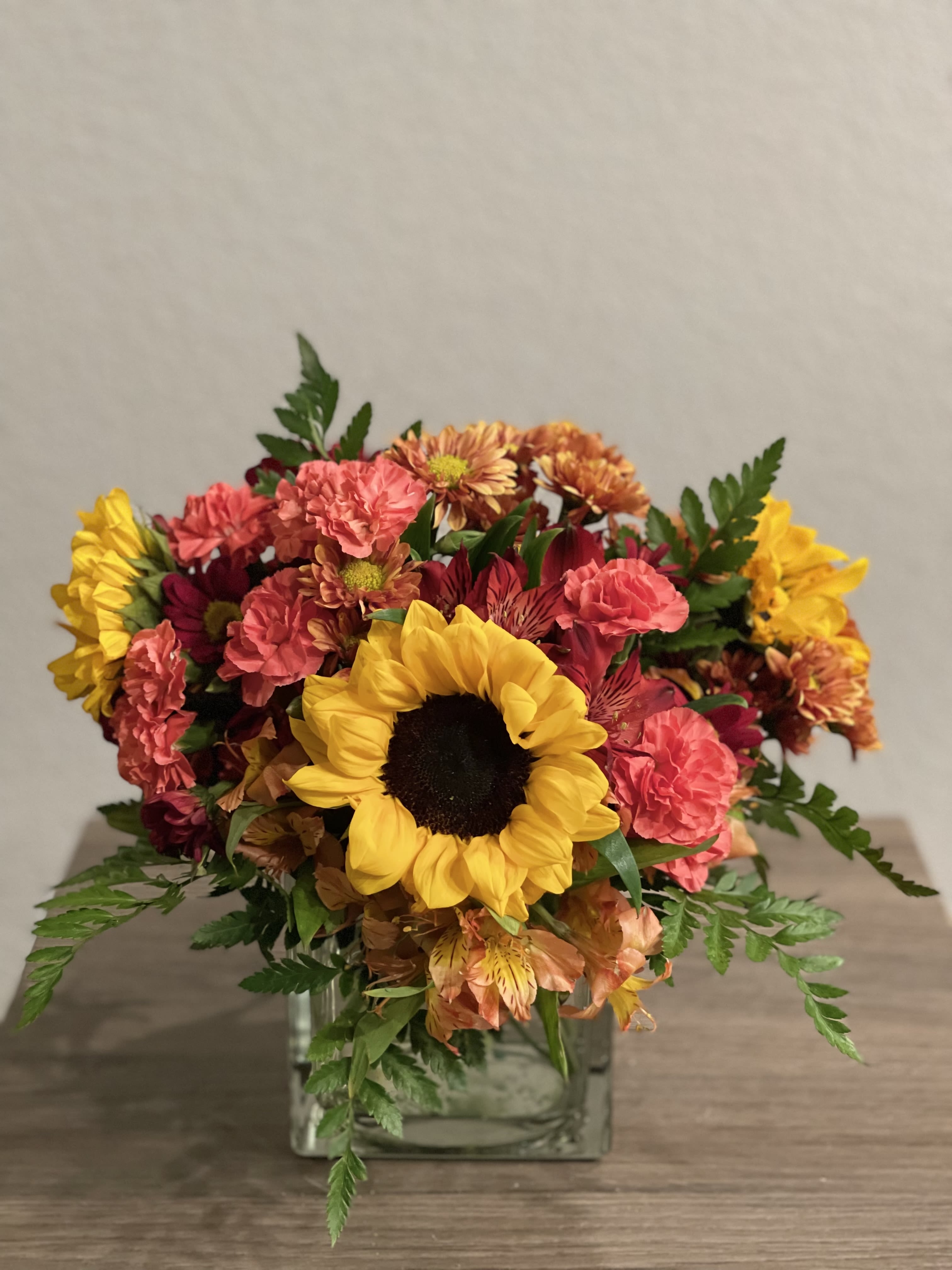 Summer Radiance Bouquet - Beautiful floral arrangement of orange carnations, sunflowers, alstroemerias flowers and leather leaves to give the final touch place on a glass square vase. .