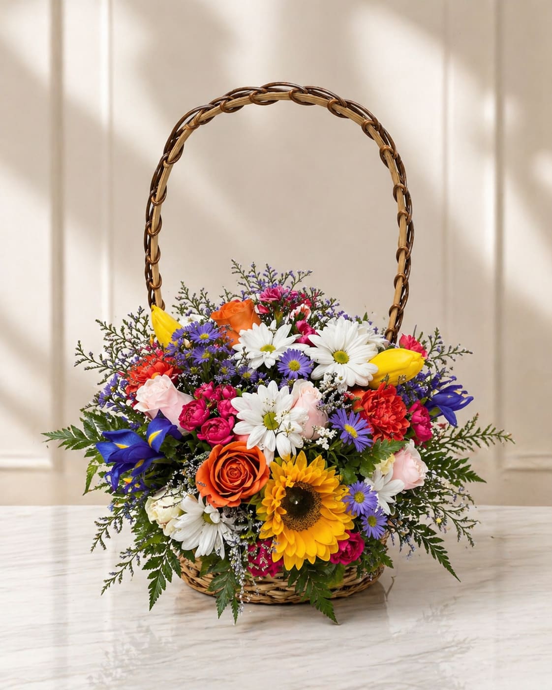 Garden mix basket - A lively, garden-style basket that reads like a sunlit patch of flowers-perfect as a birthday bouquet, congratulations gift, or a cheerful "just because" surprise. The flower arrangement in a woven basket with a tall arched handle mixes white daisies, a large yellow sunflower, pink and orange roses, yellow tulips and pops of blue and purple asters for a bright, celebratory look that will make the person you're thinking of feel genuinely seen and cheered.  This rounded, dense composition combines soft petals and textured blooms for a welcoming, abundant feel: creamy white daisies soften the bold sunflower center, while roses and carnations add romance and fullness and tulips contribute spring-fresh shape. The basket presentation makes it easy to set on an entry table, kitchen counter, or desk; the arrangement's mixed colors-white, pink, yellow and multicolor accents-work across many occasions and styles, from casual thank-you gestures to festive celebrations.