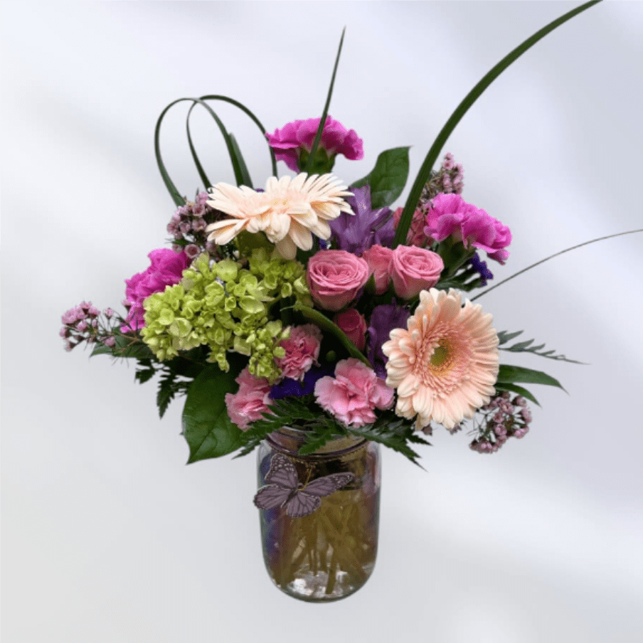 MD60 A Floral Hug - Compact and lively, this design sits in a decorative jar style glass vase with a tightly clustered bloom mass and long looping blades of foliage that rise above for an airy, sculptural silhouette. Textures range from the velvety rose petals to the bold gerbera faces and the lush hydrangea clusters, so the arrangement looks abundant from every angle. 