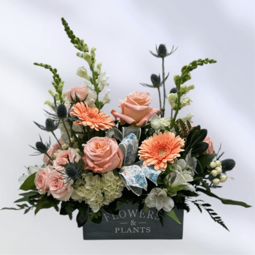 MD64 Mom's Garden - This arrangement features a soft palette of pink, peach, white, green and touches of blue. Housed in a decorative box, the design is compact at the base but airy on top, delivering a balanced, hand-gardened impression that suits spring occasions or everyday celebrations. .