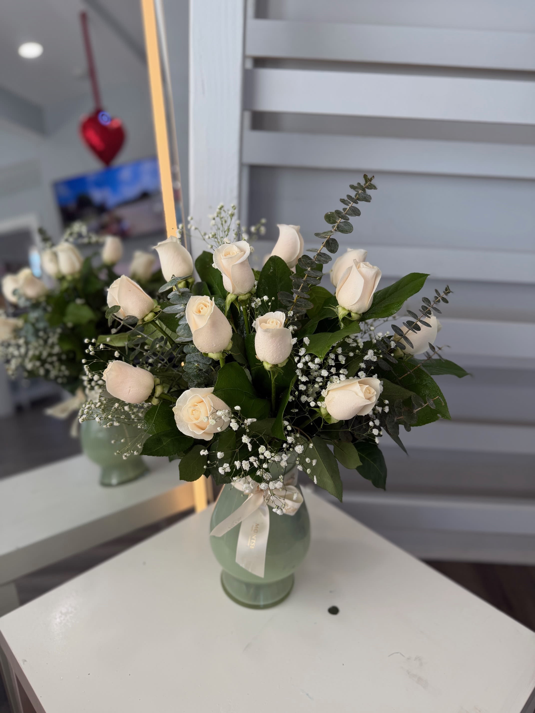 White Roses  - Rounded cluster of 12 creamy white roses with baby's breath and dark greenery in pale green vase with ribbon