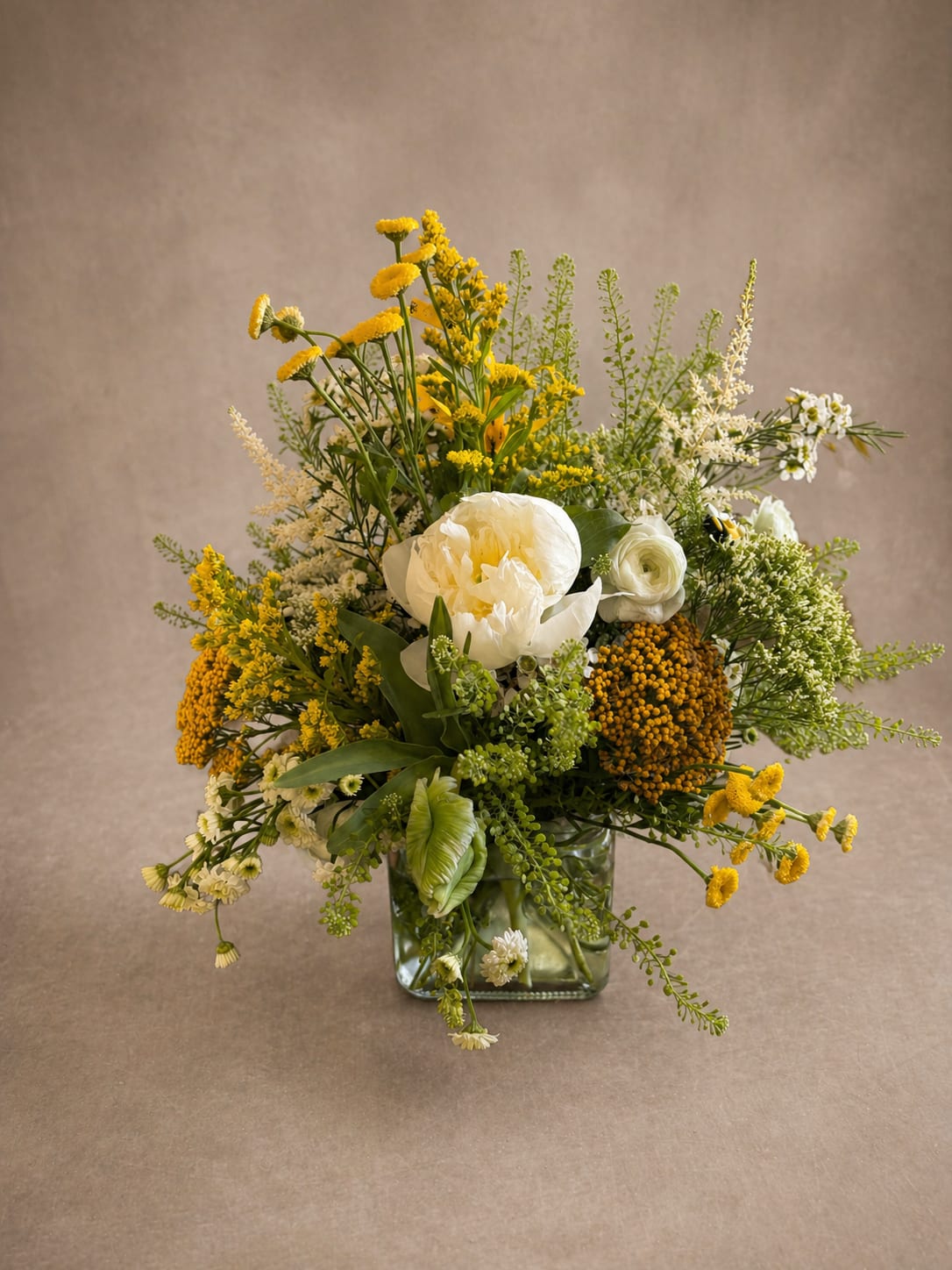sun and honey - Bright, wild, and wonderfully alive. This garden-inspired arrangement captures the feeling of a summer meadow in full bloom — golden tansy, cheerful santolini daisies, and rich goldenrod spill freely around a stunning ivory peony and delicate ranunculus, all nestled in a sleek glass cube vase. Lush greenery weaves through every stem, giving this arrangement an effortless, just-picked quality that feels both organic and artfully designed. Light enough for a baby shower, joyful enough for a birthday, elegant enough for a wedding table — Sun &amp; Honey is the arrangement that fits every room and every reason to celebrate. Because some moments don't need a special occasion — they just need flowers.