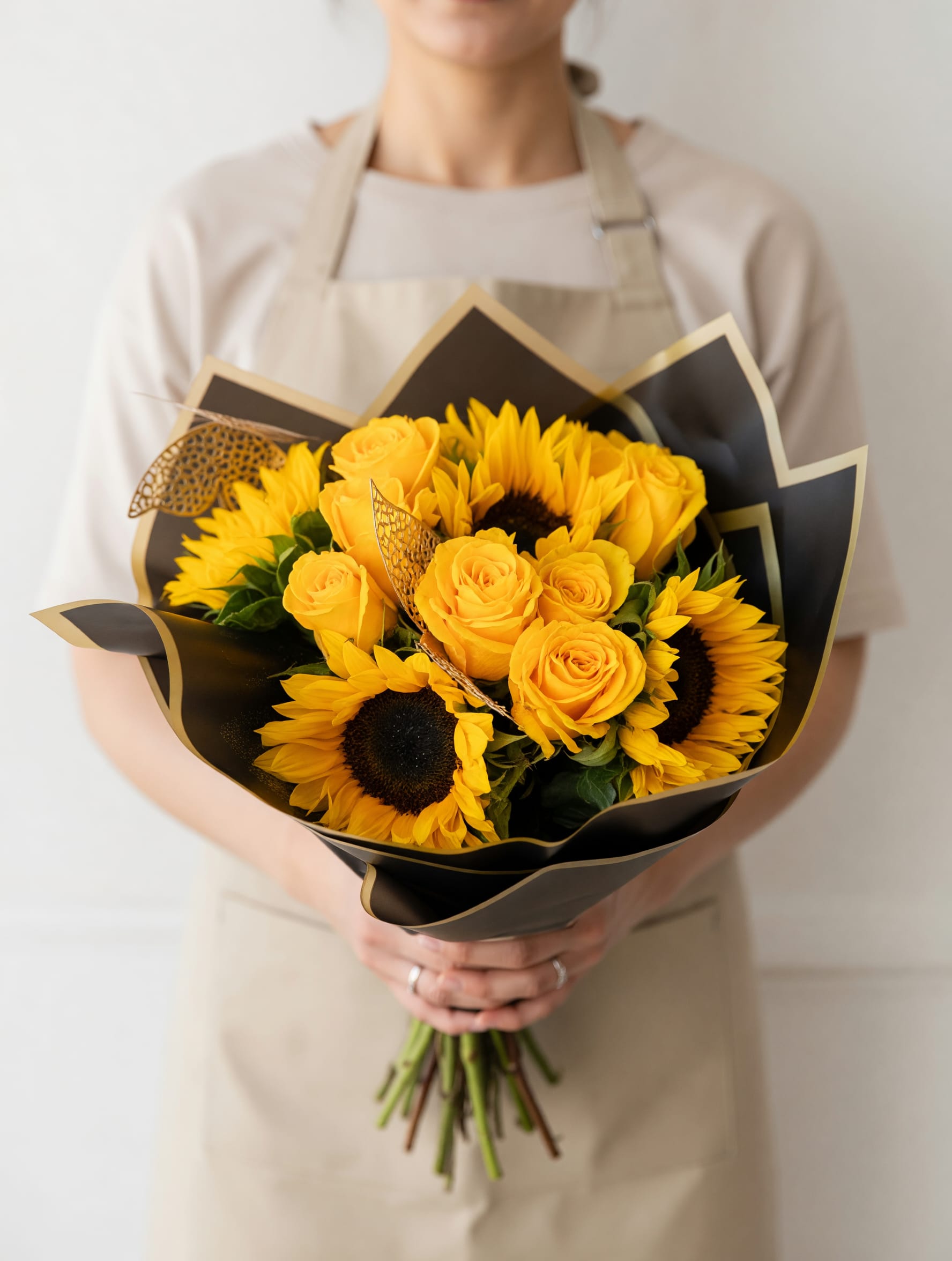 SUNNY DELIGHT - Bright yellow roses paired with cheerful sunflowers give this hand-tied flower arrangement a bold, sunny personality that instantly lifts the mood - perfect as a birthday bouquet, a congratulations gift, or a bright romantic surprise. The warm yellow blooms pop against layered matte black paper wrapped with gold trim, creating a modern, polished contrast that reads elegant and celebratory. Delicate butterfly picks add a whimsical, personal touch so the person you're thinking of will feel truly seen.  Carefully hand-tied in a rounded, hand-held shape, the arrangement includes textured accents like decorative mesh and ribbon tucked among the stems for depth and movement. The combination of roses and sunflowers offers both soft petal romance and big, joyful faces that last - a striking presentation for everyday gifting, thank-yous, or special milestones. Packaged in black floral wrap with gold edging and finished with decorative touches, it arrives ready to hand over and enjoy.