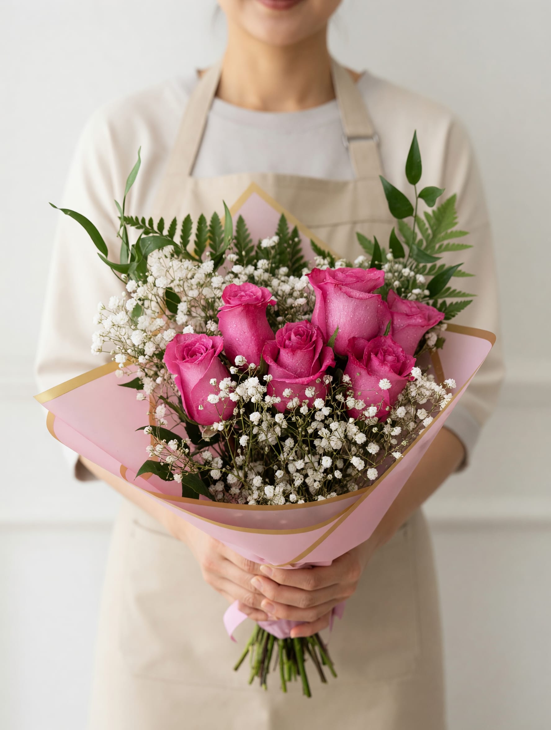 LAVENDER LOVE - Bright pink roses are clustered at the heart of this hand-tied bouquet, framed by airy white baby's breath and a mix of fresh greenery for a romantic, celebratory feel-perfect as a birthday bouquet, anniversary roses, or a Valentine's Day gift. The soft pink wrapping paper with gold trim gives the presentation a polished, feminine touch so the person you're thinking of will feel genuinely seen the moment they receive it.  This medium-sized, hand-held arrangement has a balanced, rounded shape with the roses as the clear focal point and delicate sprays of baby's breath adding texture and lightness. Wrapped in pink paper edged in gold, it arrives ready to gift and makes a sweet, classic statement for love-themed or congratulatory occasions. For best enjoyment, keep stems in fresh water and recut them before placing in a vase to extend bloom life.