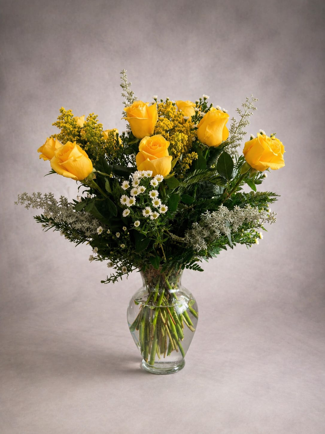 Golden Hour -  A dozen yellow roses, arranged to perfection. Radiant and full of warmth, Golden Hour is a celebration of life's brightest moments. A dozen sun-kissed yellow roses rise above a bed of delicate white aster daisies, airy goldenrod, and soft silvery foliage — all gathered in a classic glass vase that lets every stem shine. Yellow roses speak a language all their own — friendship, joy, new beginnings, and the kind of love that feels like sunshine. Whether you're celebrating a milestone, brightening someone's day, or simply saying I'm thinking of you, this arrangement says it beautifully. Some gifts don't need a reason. They are the reason.