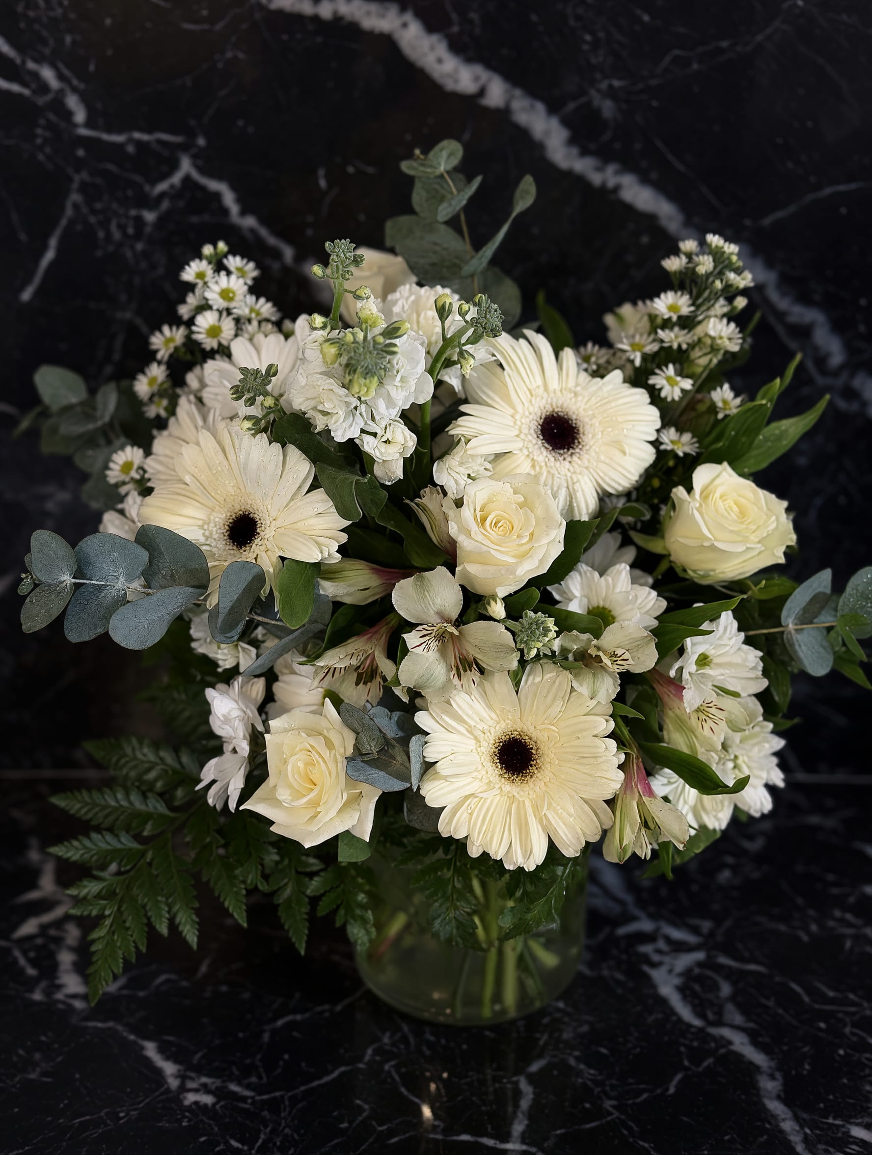 Ivory garden cube  - Creamy white gerbera daisies and ivory roses form the heart of this ivory garden cube - a rounded flower arrangement perfect as a sympathy tribute, a wedding table accent, or a refined everyday gift for someone who appreciates quiet elegance. The pale palette of white, soft pink hints, and fresh green tells the recipient you're thinking of them with gentle calmness; she'll feel comforted, seen, and quietly celebrated.  The bouquet is arranged in a clear glass cube vase and includes clusters of small daisy-like filler blooms, pale alstroemeria, stock, and cool blue-green eucalyptus for texture and contrast. Deep green foliage fills the design for a lush, compact look while the soft tones and restrained composition keep the overall impression airy and formal. This versatile arrangement arrives ready to display and suits occasions from thank-you and thinking-of-you to sympathy or spring celebrations.