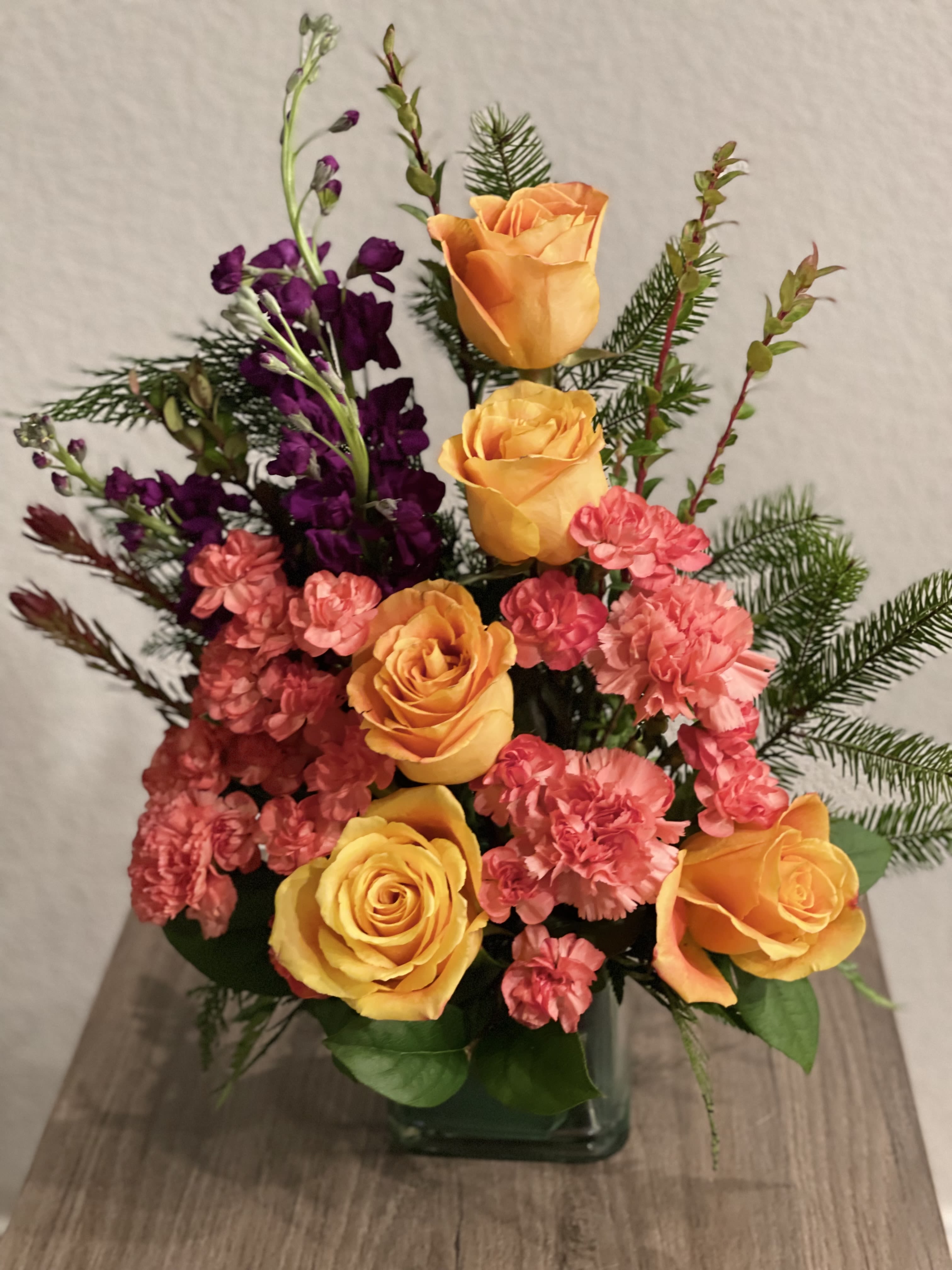 Autumn Surprise - Lovely floral arrangement of yellow roses, purple stock flowers and orange carnations and greens to give the final touch on a clear square vase. 