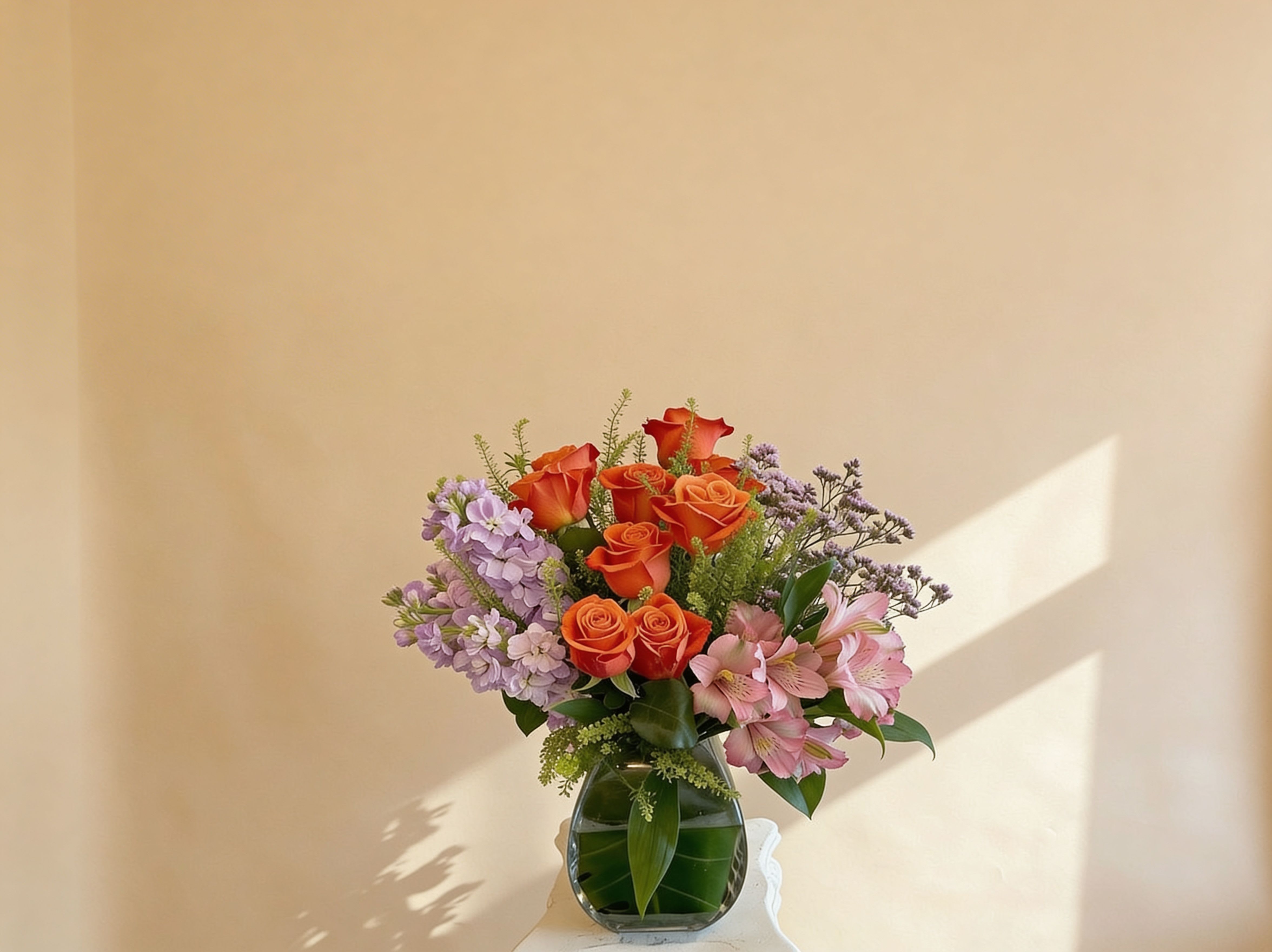 Orange roses with pink and lavender flowers in a clear glass vase on a light background