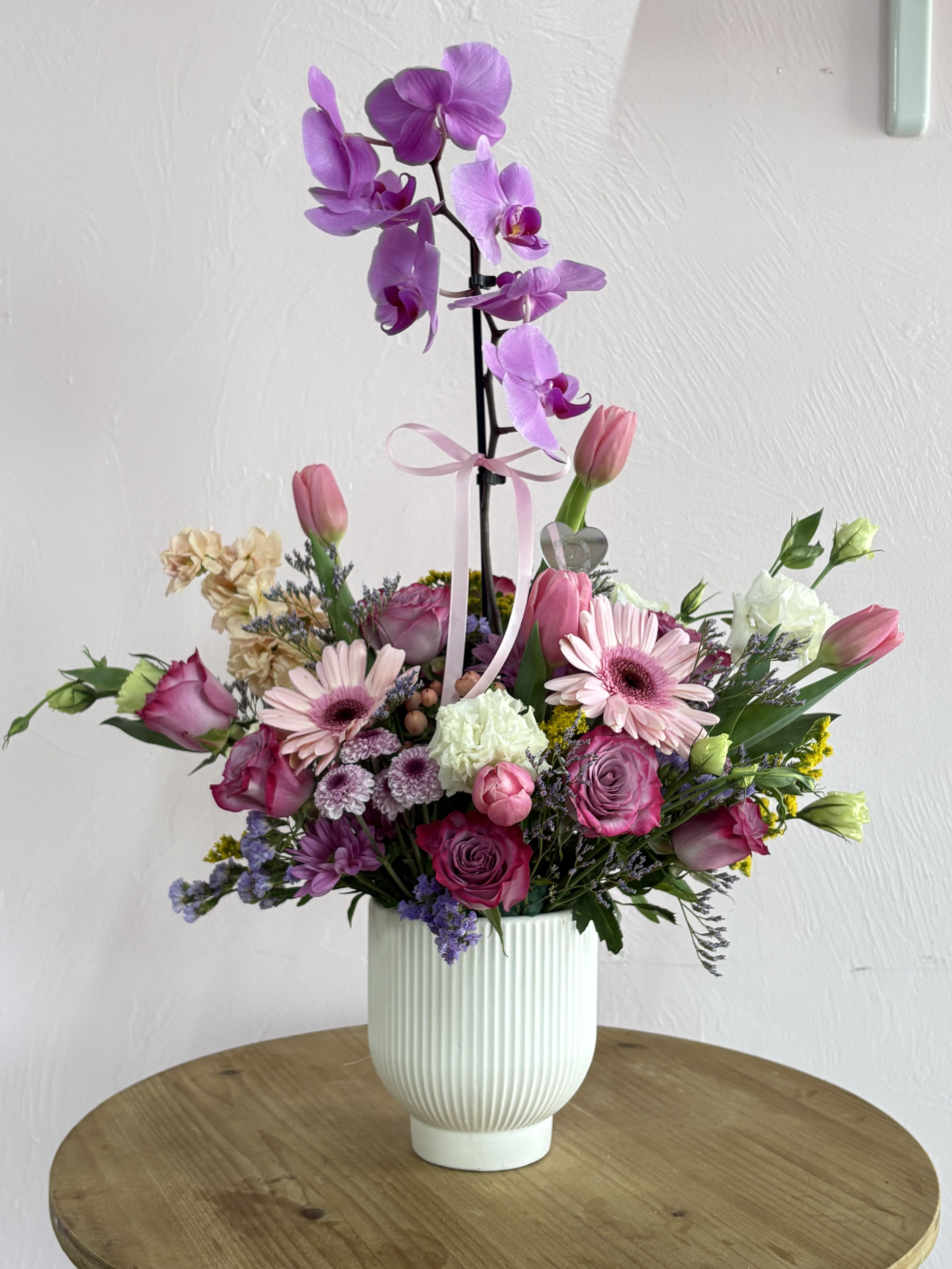 Blooming Orchid  - A graceful purple Phalaenopsis orchid stem rises above a soft pastel cloud of blooms in this romantic birthday bouquet and anniversary arrangement - the kind of gift that makes the person you're thinking of feel truly cherished. The combination of orchids, pink and lavender roses, gerbera daisies, tulips and lisianthus creates a refined, layered silhouette that reads as both elegant and affectionate, perfect for celebrating birthdays, anniversaries, or saying "I'm thinking of you."  The lower rounded cluster is dense with pink roses, soft lavender carnations and chrysanthemums, creamy-white tulips, and pops of gerbera daisies, all accented by delicate purple statice and fresh green foliage for contrast. The tall purple Phalaenopsis adds striking vertical interest and a gentle tropical scent, while a white ribbed vase, tied with a pink ribbon and finished with a heart pick, gives the whole design a polished, gift-ready presentation.  This arrangement suits anyone who appreciates classic romantic colorways and long-lasting blooms - she'll feel celebrated from the moment it's placed on her table. Vase height and the orchid stem create an airy top and fuller base; standard care like fresh water, indirect light, and keeping the orchid's roots moist will help blooms last longer. Order for same-day delivery when available to ensure the freshest presentation.