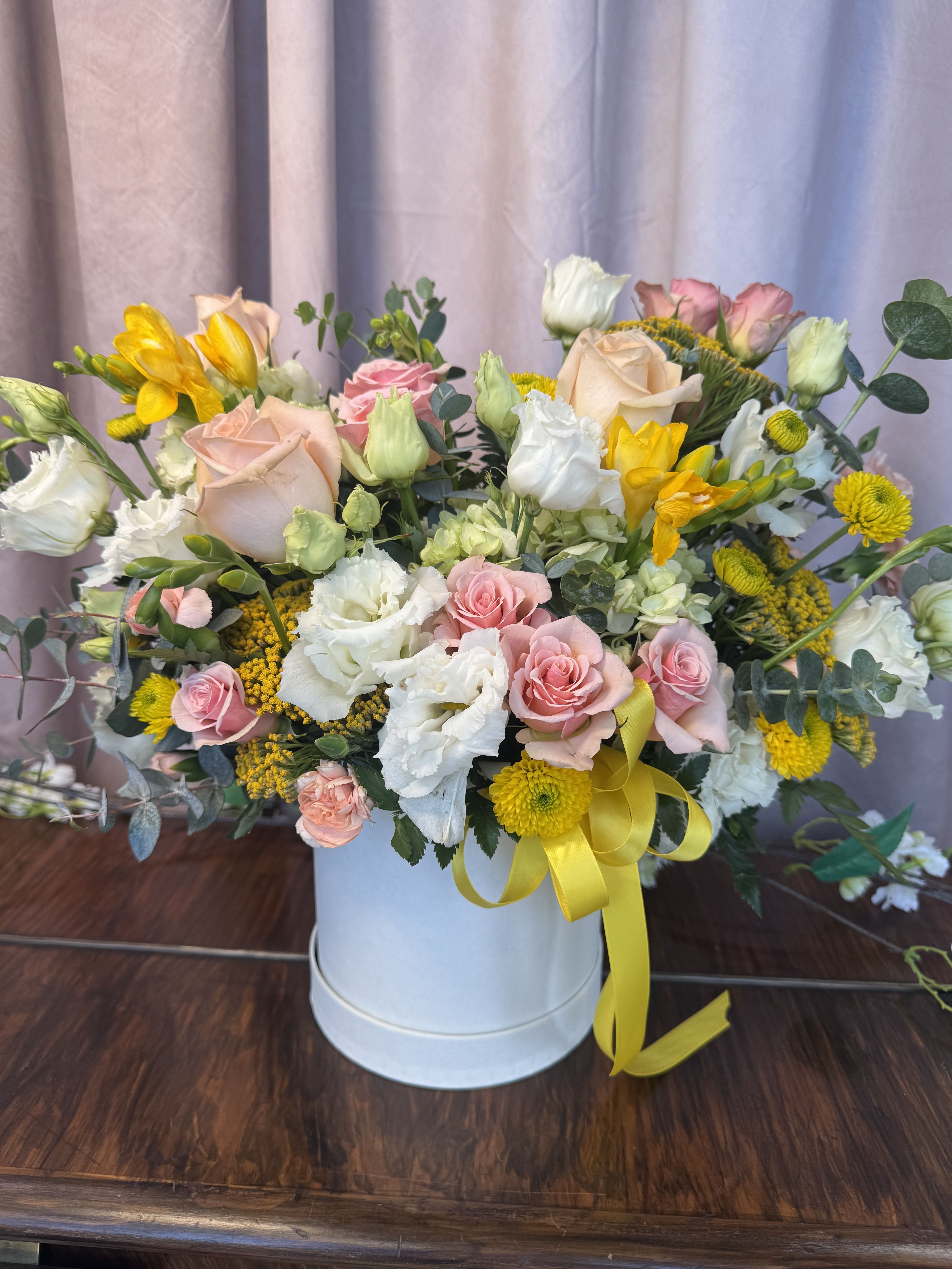 Sweet Garden Delight  - A lush, low hatbox arrangement that reads like a sunlit cottage garden - the perfect springtime flower arrangement for birthdays, thank-you gifts, or to celebrate a special moment. Sweet Garden Delight features blush pink roses and creamy white lisianthus set against cheerful yellow daisy-like blooms, creating a soft, romantic bouquet that still feels bright and joyful for occasions like Mother's Day or a get-well surprise.  Packed into a white hatbox and finished with a yellow ribbon, this mixed-color arrangement is full and textured: clustered filler blooms and silvery eucalyptus weave between the focal roses and lisianthus for gentle contrast and movement. The compact, low profile makes it ideal for tabletops or a bedside table; the hatbox provides a tidy, gift-ready presentation that keeps flowers secure during delivery.  Choose this arrangement when you want the recipient to feel warmly celebrated and seen - it's the kind of gift that looks thoughtfully arranged and instantly uplifting. Standard care: keep in a cool spot away from direct sun, refresh water if an inner water source is used, and enjoy the bouquet's soft scent and long-lasting charm throughout the season.