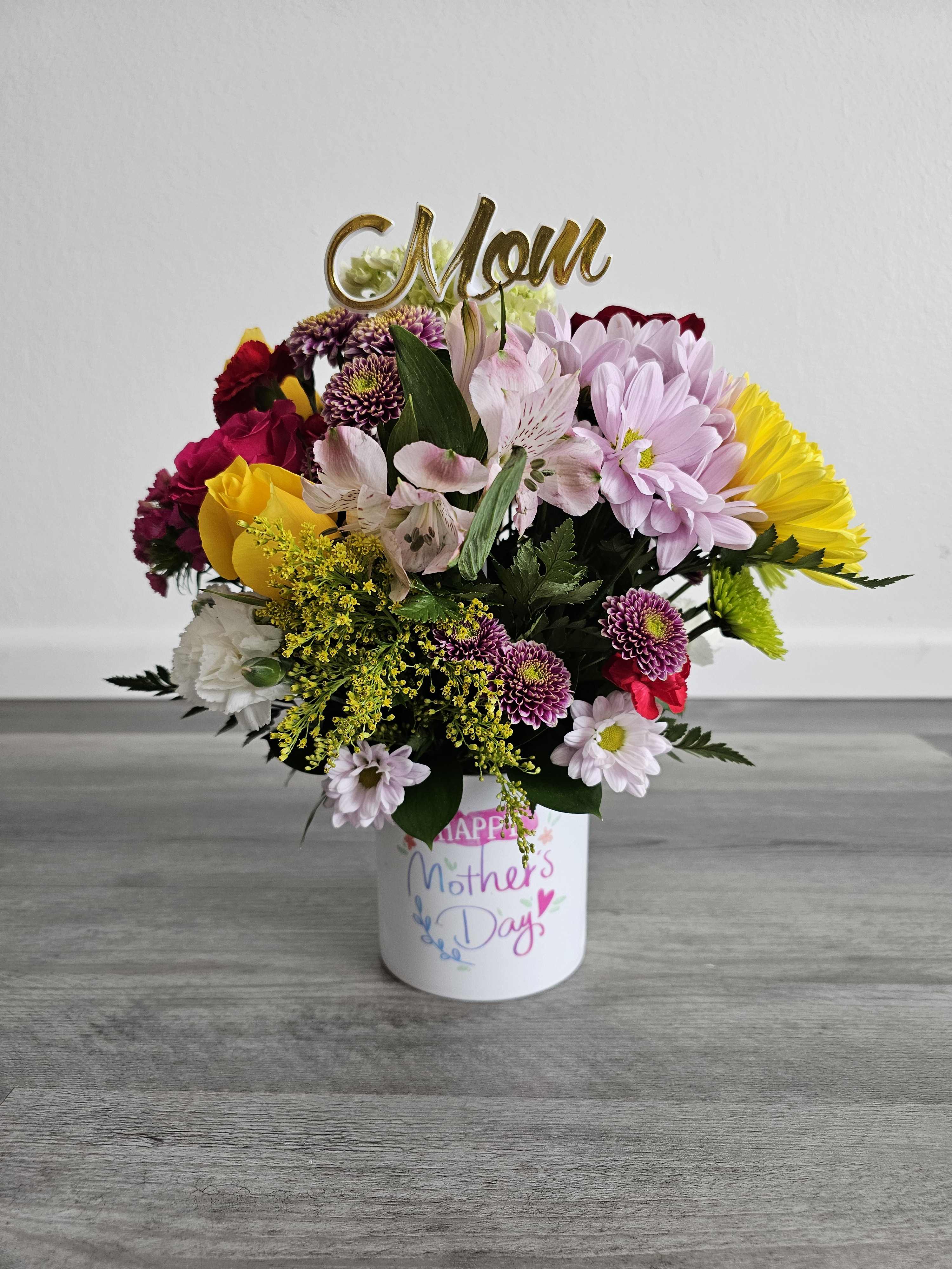 Mixed flower arrangement in a white Mother's Day container with a gold Mom topper
