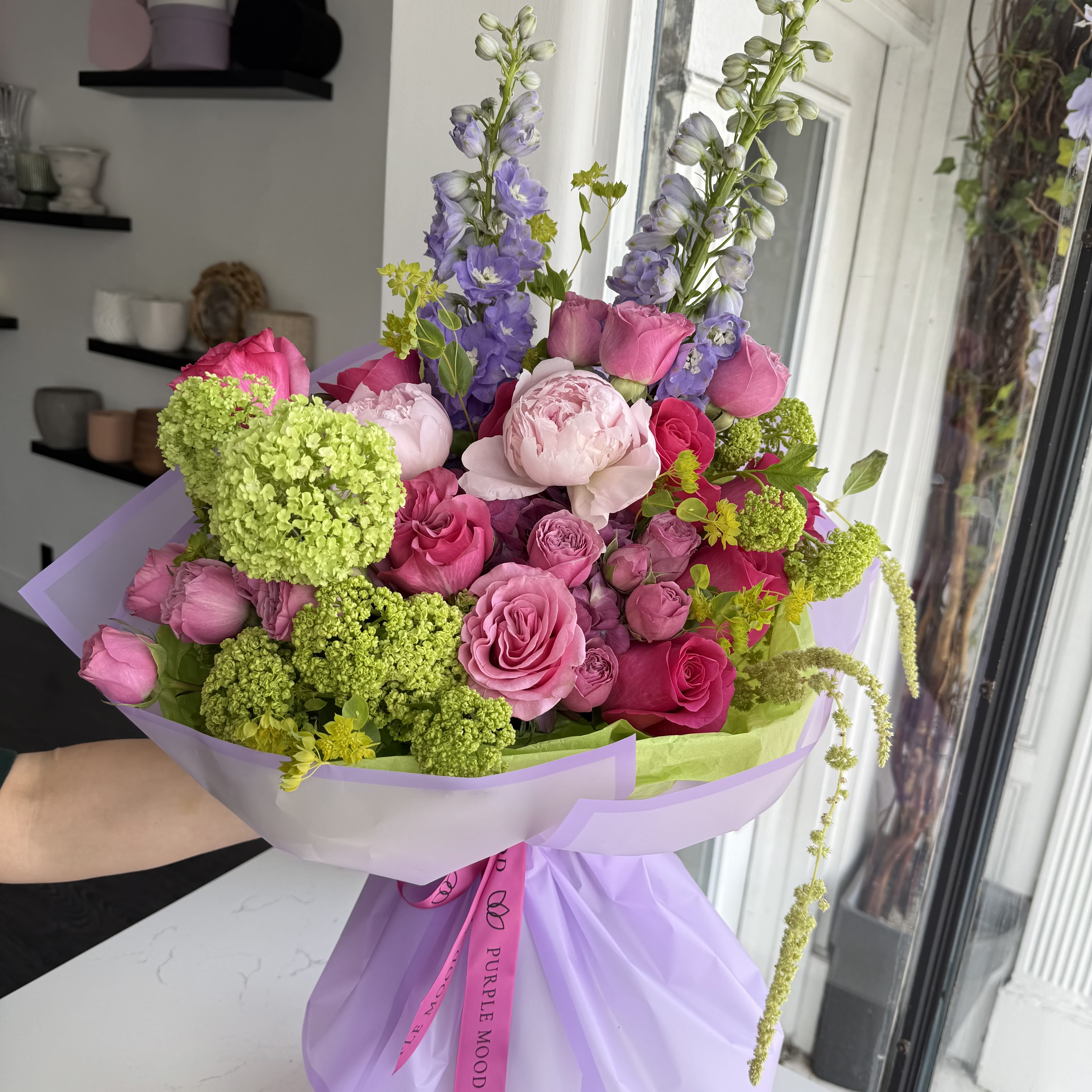Peony Kiss Bouquet - The Peony Kiss Bouquet bursts with bold pinks, soft lilacs, and fresh greenery, creating a playful yet romantic design. With its mix of vivid tones and lush peonies, it’s like a kiss captured in flowers — bright, passionate, and unforgettable.      •    Romantic mix of pink blooms with a touch of lilac and green     •    Wrapped in elegant lilac paper with signature ribbon     •    Perfect for love notes, birthdays, or any heartfelt surprise  VASE NOT INCLUDED