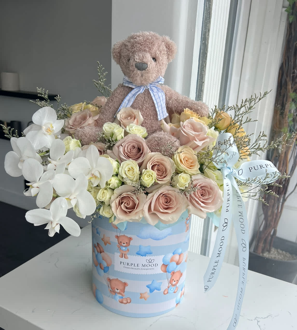 Teddy Bear Box - All around box.   Standard container size - D8.25'x H9.5'  Deluxe container size - D10.5'x H12.25'  Teddy Bear Box A sweet and heartwarming gift, the Teddy Bear Box combines soft pastel blooms with an adorable plush teddy, making it perfect for welcoming a new baby, celebrating a birthday, or surprising someone special. This arrangement is both elegant and playful, guaranteed to bring smiles and warm memories.      •    Includes a soft teddy bear for an extra touch of love     •    Pastel-toned arrangement in a themed keepsake box     •    Ideal for baby showers, birthdays, or “just because” moments     •    A beautiful mix of charm, comfort, and elegance