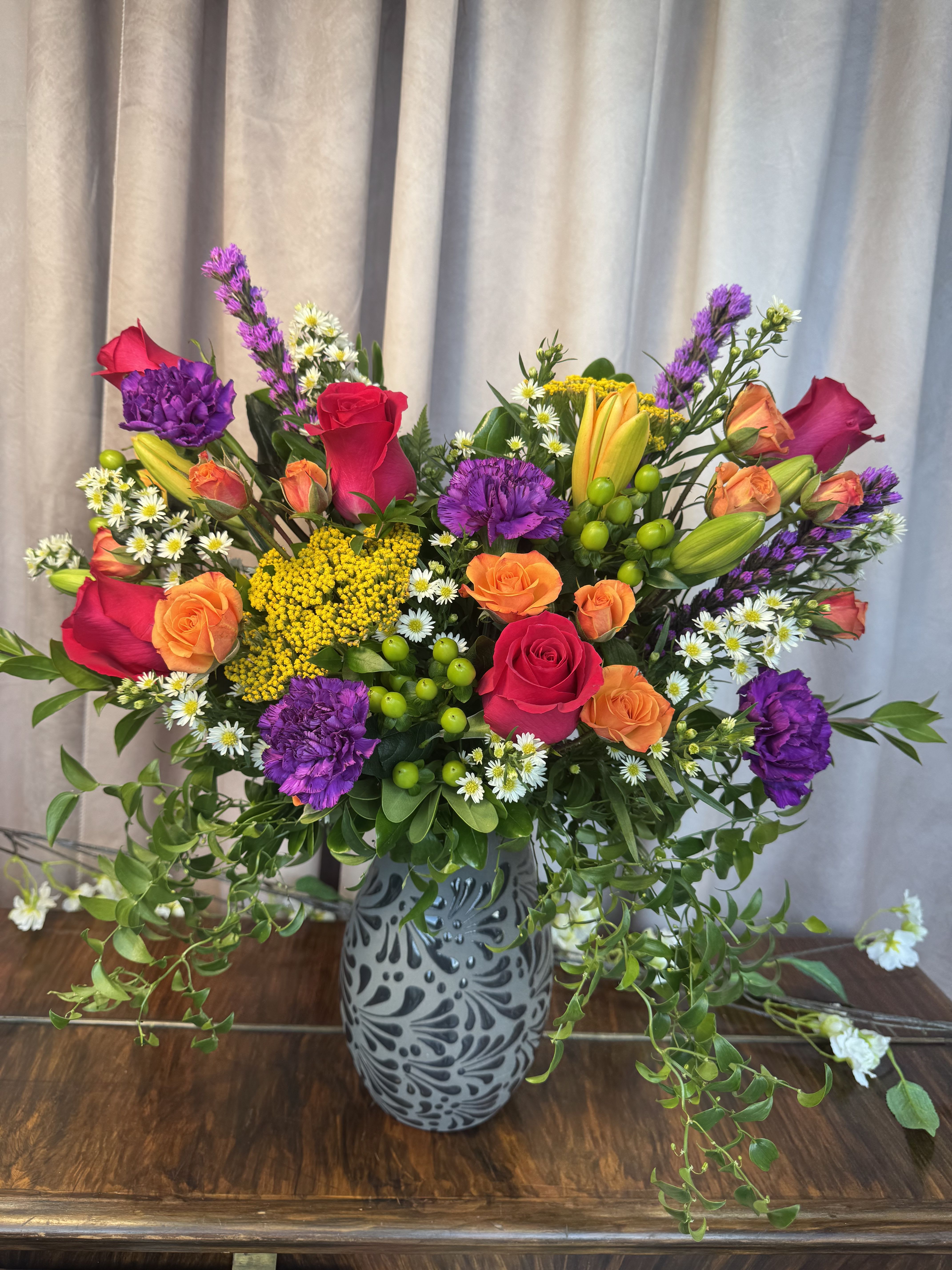 Encanto Floral  - A lively multicolor bouquet that feels like a celebration in bloom - bright pink and orange roses paired with purple carnations and creamy lily buds create an uplifting birthday bouquet or congratulations arrangement that will make the recipient feel truly celebrated. The mix of small white daisy-like blooms and clustered yellow filler adds cheerful contrast, while the loose, airy design gives the composition a fresh, spontaneous look perfect for sending to a friend, coworker, or family member.  Arranged in a patterned gray ceramic vase, this large, full display measures as a generous centerpiece with trailing greenery that extends beyond the vase for extra presence and visual movement. The combination of roses, carnations, lilies, daisies and assorted filler offers varied textures and blooms at different stages - buds and open flowers together - so the bouquet keeps opening and delighting over several days.  Ideal for birthdays, congratulations, or "just because" moments when you want someone to feel noticed, this arrangement is designed to arrive ready-to-display in its ceramic vessel. To keep blooms looking their best, remove any foliage below the waterline and change the water every few days; the mix of sturdy roses and carnations with longer-lasting lilies and daisies gives the recipient staying power and continuous color.