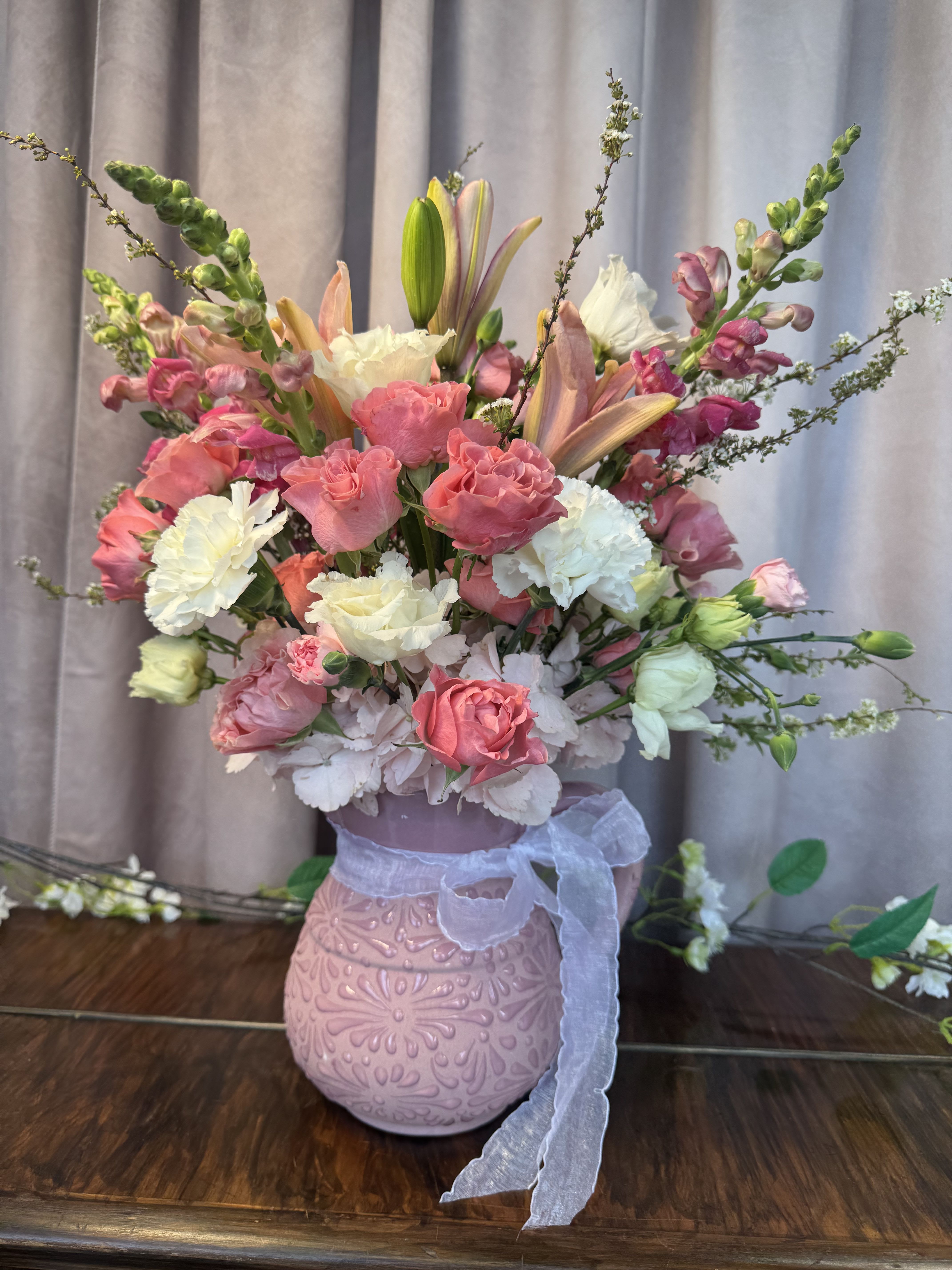 Blooming Pink  - A lush, romantic display of pink and white blooms arranged with airy movement - this flower arrangement is a perfect birthday bouquet or spring celebration gift that feels both feminine and refined. The mix of roses, ruffled carnations, and tall snapdragon spikes creates a gently layered silhouette, while a few lily buds add anticipation and soft fragrance that the recipient will notice as they open the room.  Set in a decorative pink ceramic vase and tied with a sheer ribbon, the composition balances full, garden-style roses and carnations with vertical snapdragons for height and visual drama. Hints of green foliage and touches of purple among the blooms add depth to the pastel palette, and the lily buds promise a gradual opening over the next few days.  Give this to someone you want to make feel truly celebrated - it's ideal for anniversaries, bridal showers, Mother's Day, or as an elegant get-well gift. Vase-ready on arrival; trim stems and change the water every two days to extend the life of the arrangement and enjoy the lilies as they bloom.