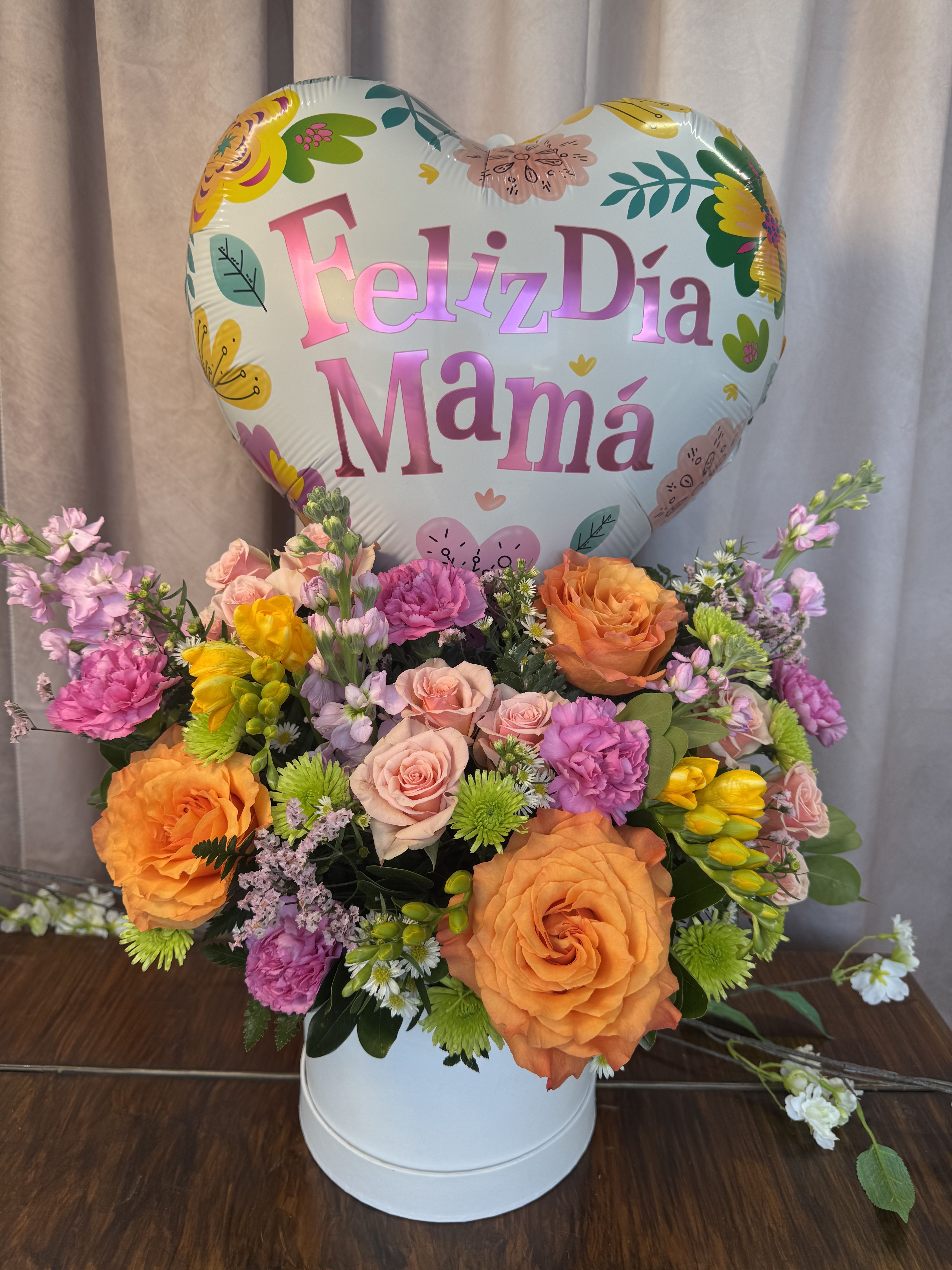Mom In Bloom  - Bright, cheerful blooms in a rounded white hat box make this flower arrangement a joyful way to celebrate Mother's Day or say "I'm thinking of you" to the mom in your life. The Mom In Bloom arrangement pairs large orange roses with smaller pink roses, soft carnations, and purple stock for a full, romantic silhouette; a heart-shaped foil balloon reading "Feliz Día Mamá" gives it an unmistakable celebratory touch that will make her feel noticed and loved.  This compact yet abundant design mixes yellow and daisy-like filler chrysanthemums for pops of sunny contrast and textured interest throughout the dense, rounded bouquet. The white hat box keeps the presentation clean and gift-ready while the varied bloom sizes create a lush, hand-tied appearance-perfect for a breakfast surprise, brunch table centerpiece, or bedside delivery that brightens her day.  Made to arrive ready-to-gift, Mom In Bloom is ideal for Mother's Day, a birthday, or any moment when you want Mom to feel truly celebrated; the balloon and hat box make assembly effortless. Choose this arrangement when you want her to open a present that reads joyful, festive, and distinctly thoughtful.