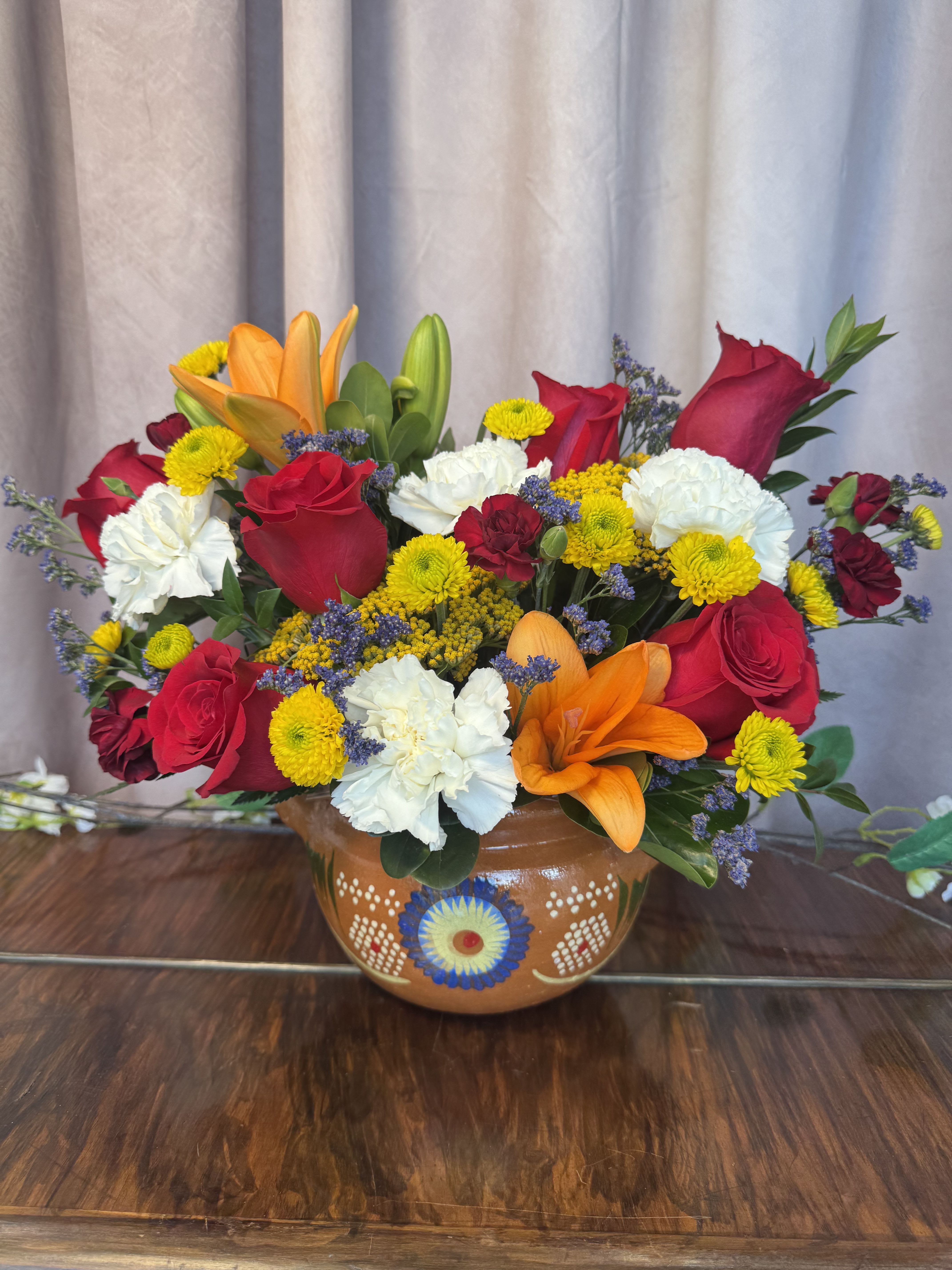 ollita de barro  - Bright red roses and vibrant orange lilies make a bold first impression in this cheerful flower arrangement-perfect as a Mother's Day bouquet or a festive birthday gift. The rounded ceramic vase gives a handcrafted, painted look that feels warm and grounded, while pops of white carnations and sunny yellow chrysanthemums brighten the composition to convey joy and appreciation to the person you're celebrating.  Textured purple filler flowers weave through the mix to add contrast and depth, giving the design a layered, garden-fresh feel. The arrangement combines roses, lilies, carnations, chrysanthemums and other seasonal blooms in a compact, rounded silhouette that looks lovely on a kitchen table or bedside dresser. Arrives ready to display in its painted ceramic vase; simple care-fresh water and a cool spot-will keep the blooms looking their best for days.