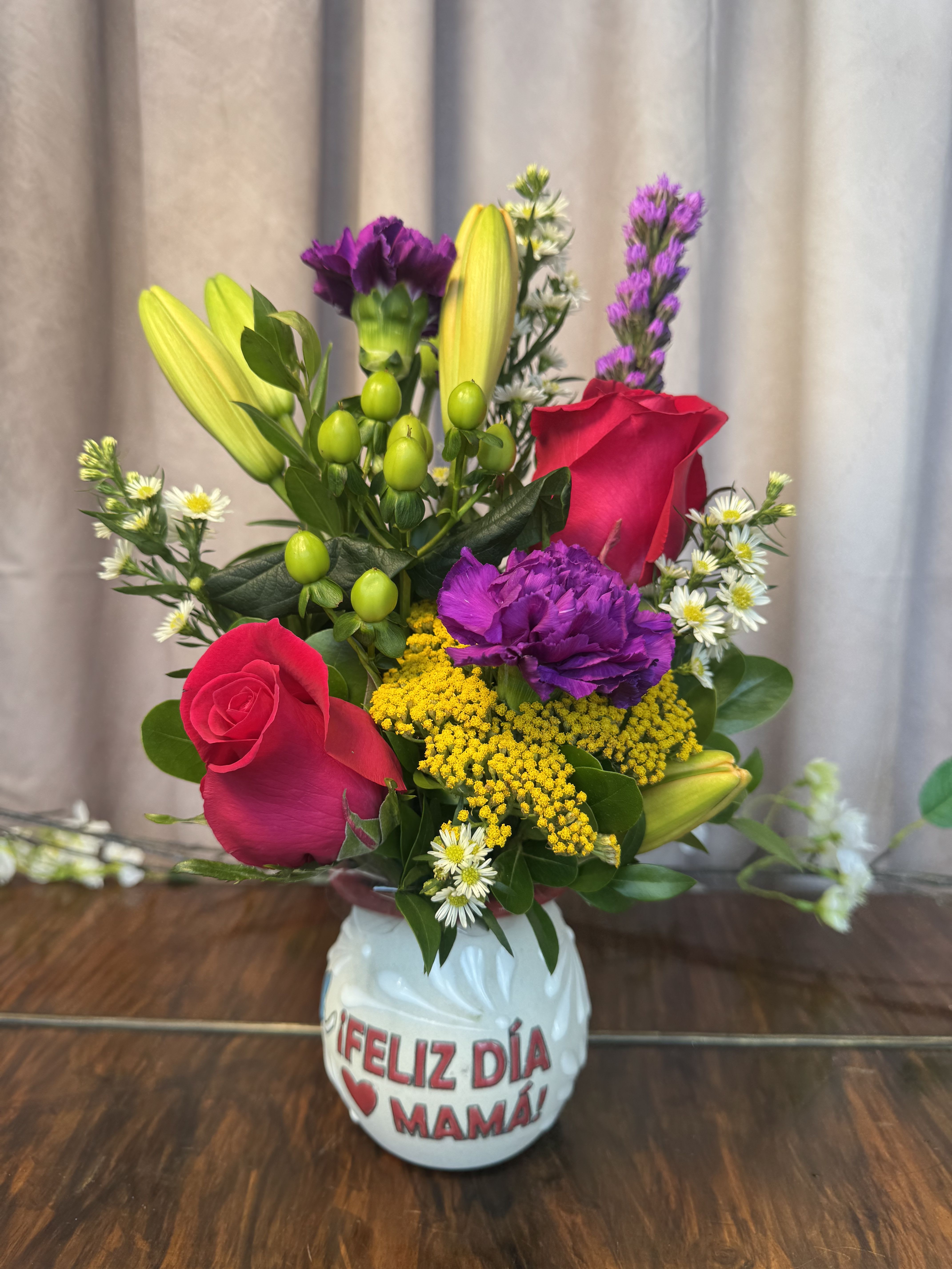 encanto floral mini - Two bright pink roses center this cheerful Mother's Day flower arrangement, a compact flower arrangement that feels like a sunny celebration for Mom. Designed as a Mother's Day bouquet, the "encanto floral mini" pairs vertical yellow lily buds and purple carnation spikes with small white daisy-like blooms and clusters of yellow filler to create lively movement and a joyful, colorful palette.  Arranged in a white ceramic vase printed with "Feliz Día Mamá," the composition balances taller lilies with fuller roses and carnations near the base for a pleasing, compact silhouette that fits on a kitchen table or bedside dresser. Colors include pink, purple, yellow, and white; flowers used are roses, lilies, carnations, daisies, and assorted filler blooms. The vase is part of the gift, ready to display-no additional container needed.  Perfect for making Mom feel celebrated and seen, this arrangement sends a message of warmth and appreciation on Mother's Day or any moment you want to say "gracias" with flowers. For best enjoyment, keep in a cool spot away from direct sun and refresh the water every couple of days to extend vase life.
