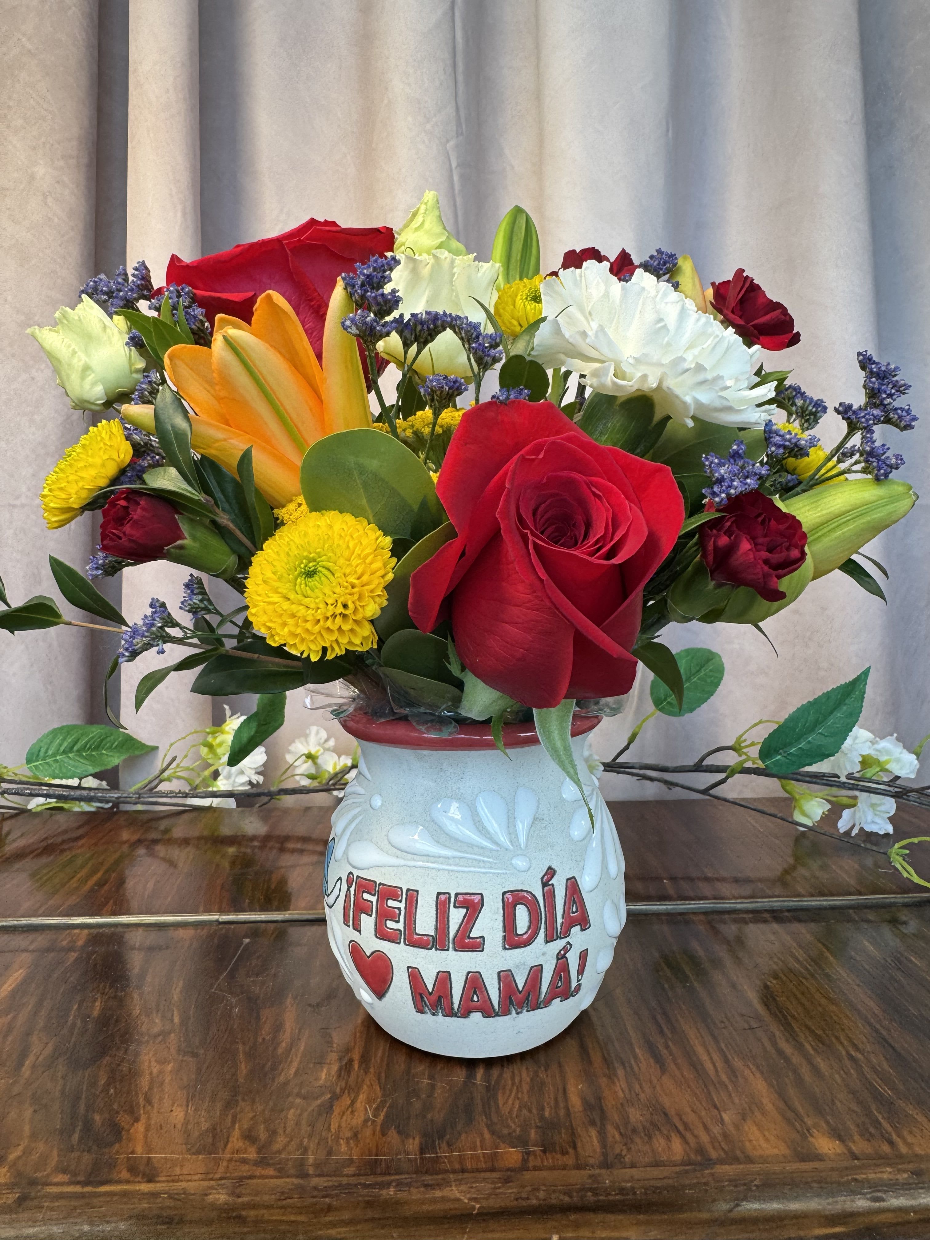 tazita de barro - Bright, celebratory color takes center stage in this Mother's Day flower arrangement-perfect as a Mother's Day bouquet or a cheerful birthday surprise for the person you want to honor. The rounded, tightly gathered design features red roses and orange-tinted lilies that read like warm, loving accents, balanced by sunny yellow chrysanthemums and crisp white blooms; small purple filler flowers add playful contrast against the warmer tones. The ceramic mug printed with "FELIZ DÍA MAMÁ!" and a heart makes the recipient feel celebrated the moment it arrives.  Arranged in a decorative ceramic tazita de barro, this compact, full centerpiece reads as a joyful yet personal gift rather than a formal statement piece. The dense mix of roses, lilies, chrysanthemums and assorted filler blooms creates a lush texture and a lively color contrast that brightens a kitchen table, bedside, or office desk. Order with confidence for Mother's Day or any occasion when you want her to feel seen-arrives ready to display in its printed vase and best enjoyed with fresh water changes every few days to keep petals vibrant.