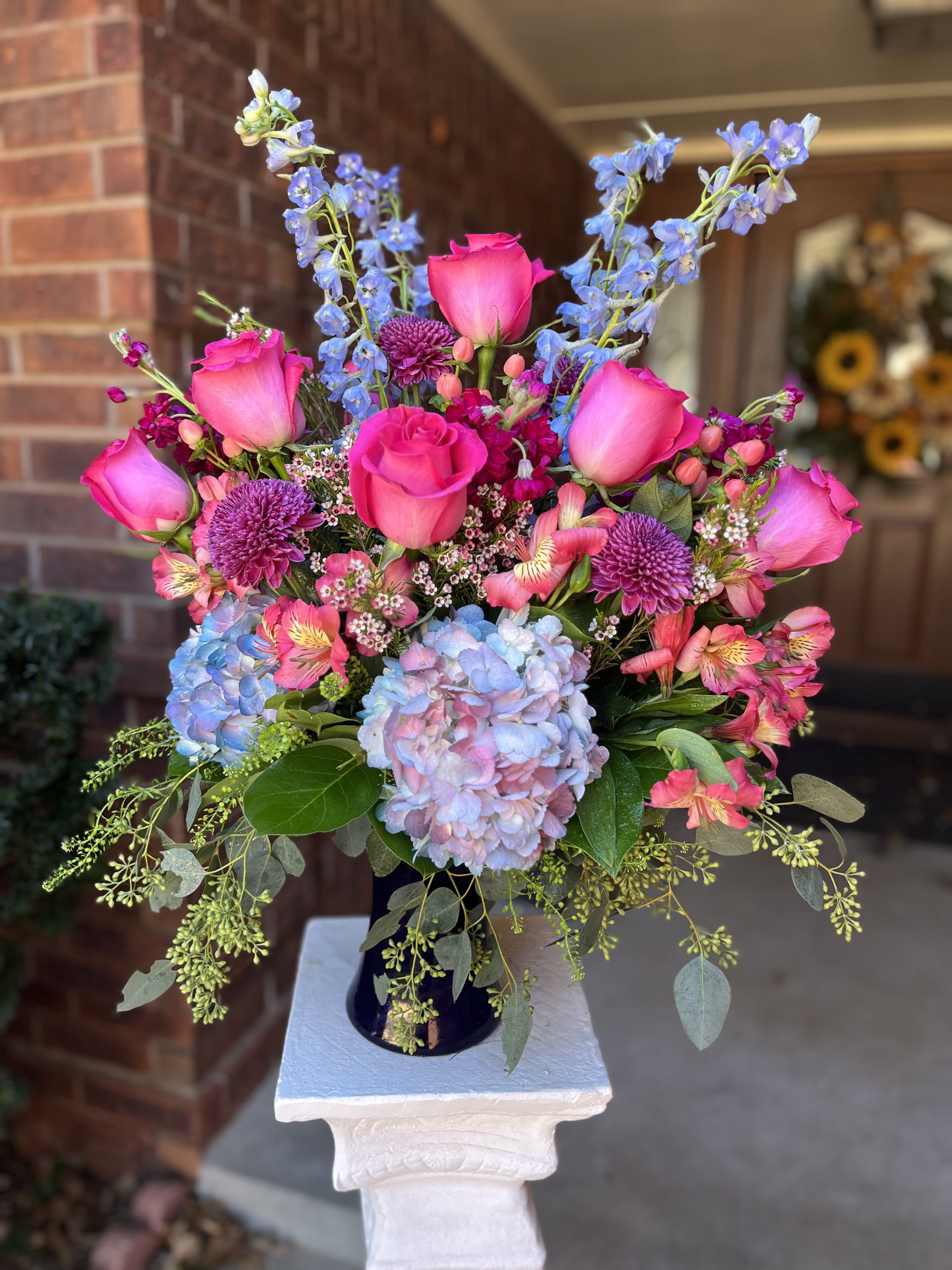 Sophia’s Rose - Vivid hot-pink roses take center stage in this celebratory flower arrangement, paired with tall blue delphinium spikes and large pastel blue-lavender hydrangea blooms for a lush, upright display-perfect as a birthday bouquet, anniversary arrangement, or anything to mark a joyful milestone. The mix of pink, blue, purple, and white creates a romantic, cheerful impression that will make the person you're thinking of feel truly celebrated.  The design includes roses, delphinium, hydrangeas, alstroemeria, dianthus, purple button mums, and small white filler blooms, with berry-like accents woven through the sides for added texture. Presented in a dark vase, the layered composition reads polished and abundant, combining tall vertical elements with full, rounded heads for visual depth. This is the kind of arrangement that arrives looking composed and ready to brighten a table, mantel, or office.