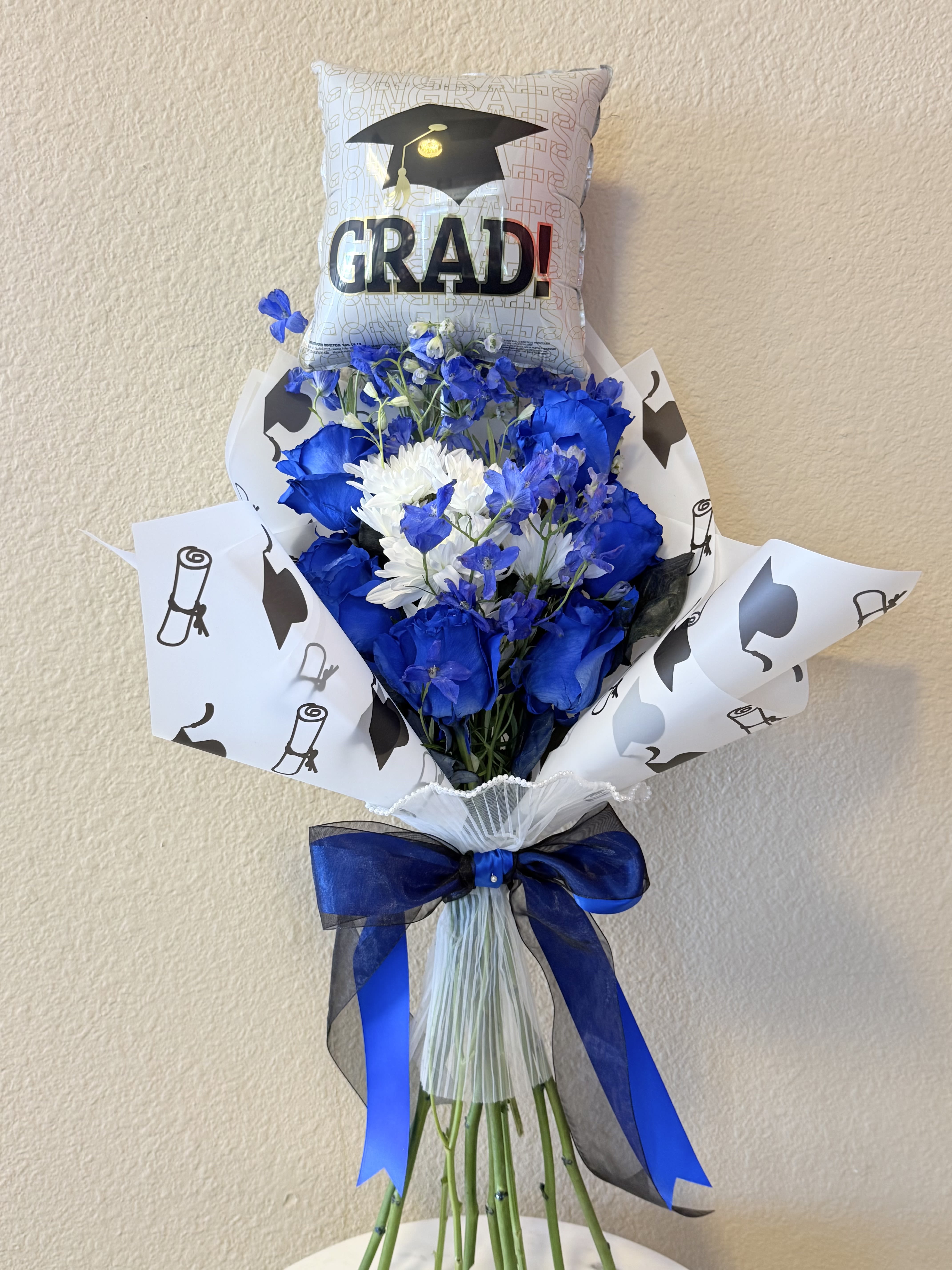 Blue Legacy Graduations Bouquet  - Vivid blue roses set a bold, celebratory tone in this graduation bouquet-perfect as a graduation bouquet, birthday surprise, or a heartfelt thank-you for the grad in your life. The striking blue-and-white color story, paired with black-accented wrapping, reads as school-spirited and joyful so the recipient will feel proudly recognized the moment they see it.  Crisp white chrysanthemums and daisy-like blooms mingle with delphinium and delicate blue filler flowers for texture and contrast, arranged in decorative white wrapping paper printed with graduation icons. A large "GRAD!" balloon and a coordinating graduation pillow rise above the stems, finished with blue-and-black satin ribbon for a polished, photo-ready presentation.  Send this bouquet to celebrate a commencement, say thanks, or mark a birthday with something memorable-it's the kind of gift that makes someone feel truly seen and celebrated. Sizes and specific flower counts may vary by availability; stems are arranged for lasting freshness, and simple water-and-vase care will keep the flowers looking their best through the celebrations.