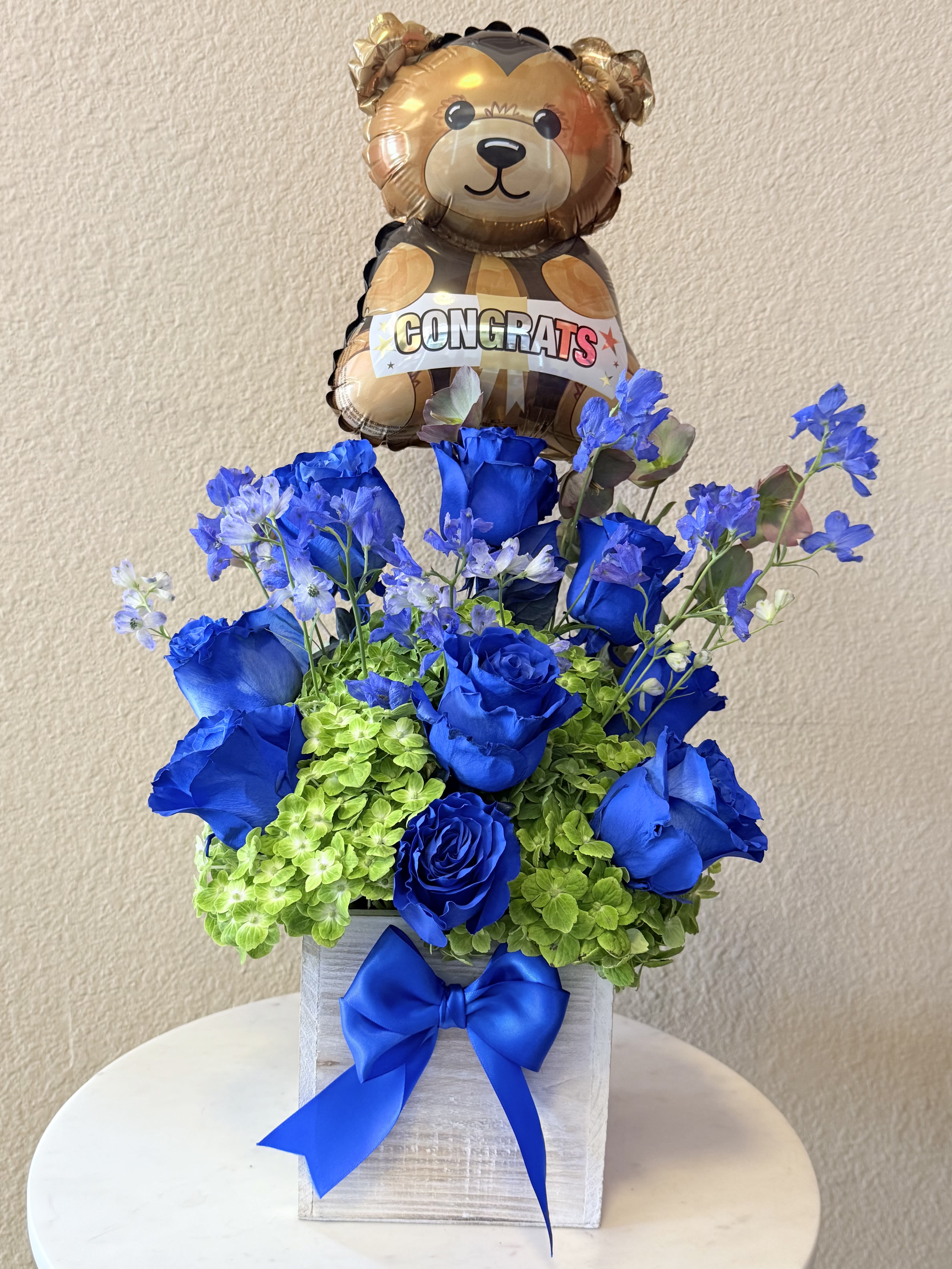 Blooming Success  - Vivid blue roses paired with bright green hydrangeas create an energetic, high-contrast bouquet that's perfect as a congratulations arrangement or a festive birthday gift. The celebratory foil teddy bear balloon marked "CONGRATS" lifts the design, making the recipient feel recognized and celebrated the moment it arrives.  Clusters of deep blue roses nestle against lush, lime-green hydrangea heads while small pale blue filler flowers add lightness and vertical texture above the denser blooms. The composition is arranged in a whitewashed decorative box tied with a large satin blue ribbon for a polished, gift-ready presentation that's easy to display.  Send this piece to mark a promotion, graduation, new baby celebration, or milestone birthday-the playful balloon and bold color story say "well done" in an unmistakable way. The boxed design arrives ready to enjoy; keep blooms in a cool spot away from direct sun and refresh the water as needed to extend their freshness.