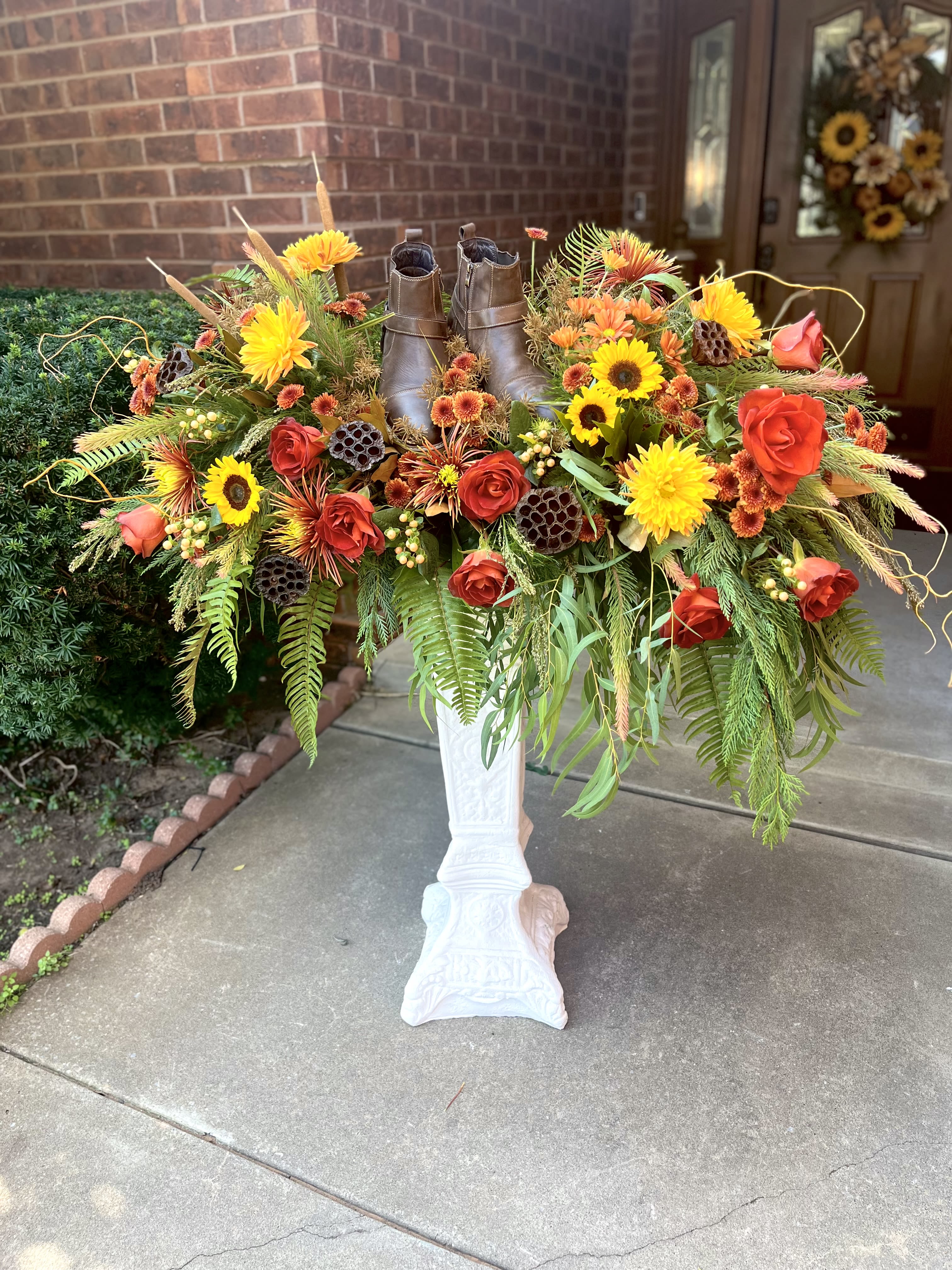 Woodland Meadows - A pair of well-worn brown boots planted at the heart of this harvest-inspired flower arrangement makes Woodland Meadows an unforgettable sympathy display to honor your loved one.   Bright yellow sunflowers dominate the composition, accented by orange and red roses and clusters of small chrysanthemum blooms in complementary tones; cascading greenery and dried seed pods add movement and rustic texture. The flowers spill from and around the boots, creating an overflowing, decorative silhouette that's casual rather than formal.   Boots not included. 