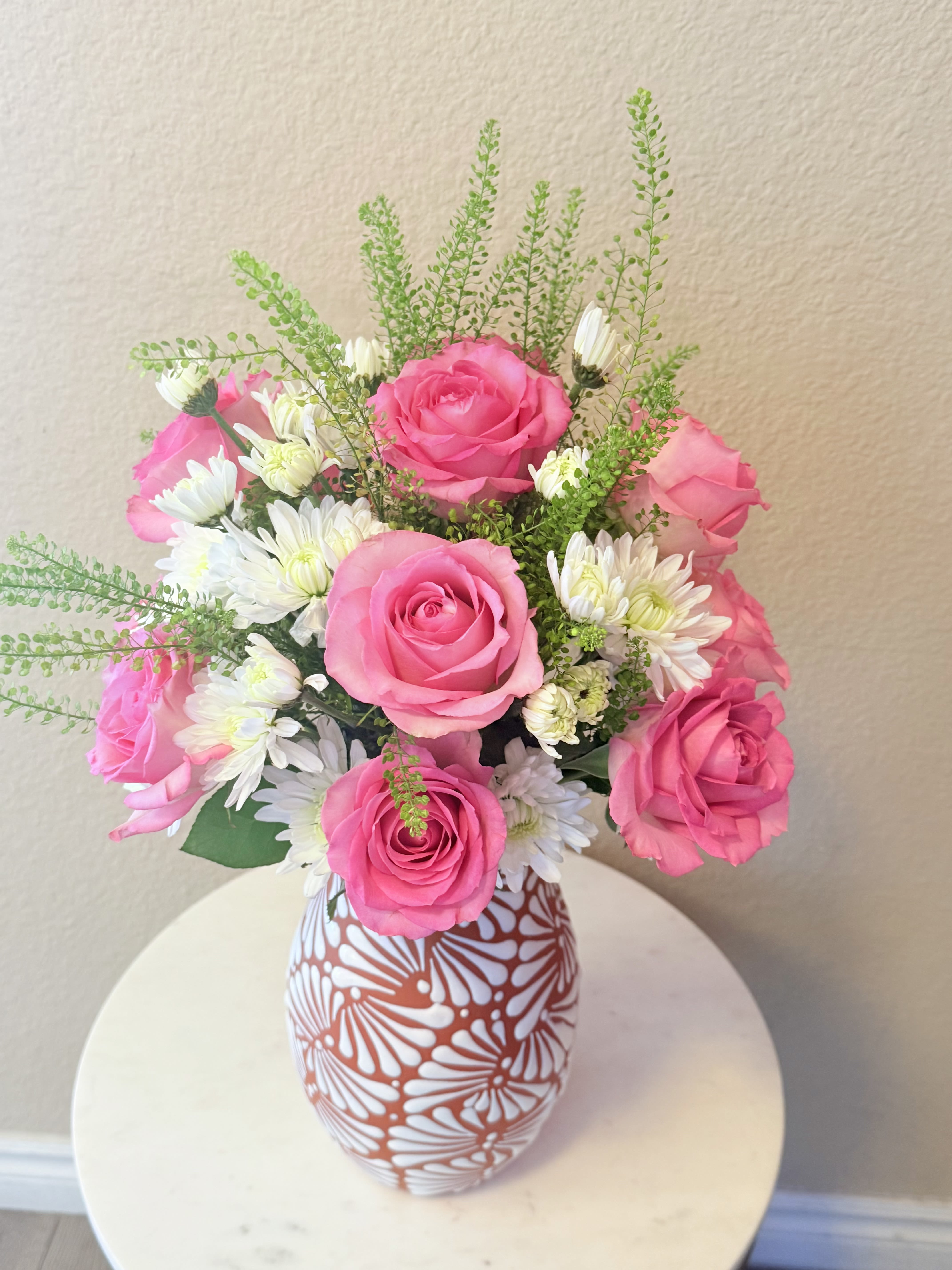 Blush Love for Mom - Bright pink roses mingle with cheerful white daisy-like blooms and airy green filler to create a joyful, romantic bouquet-perfect as a birthday bouquet, thank-you gift, or an everyday arrangement to show Mom you care. The rounded composition reads lively and affectionate; the recipient will feel celebrated and warmly thought of the moment the vase arrives.  This flower arrangement features vivid pink roses, crisp white daisies, and soft green accents gathered into a full, domed shape. It's presented in a tall ceramic vase with a bold white floral pattern against a terracotta background, adding a handcrafted, decorative touch that complements the blooms' fresh color contrast. The mix of textures-from velvety rose petals to delicate daisy centers and feathery greenery-creates visual depth and a softly romantic feel.  Ideal for Mother's Day, a birthday, or "just because" moments, Blush Love for Mom sends a message of appreciation and affection without being over-the-top. The ceramic vase makes this an easy display piece for a kitchen table, entryway, or bedside-no additional container needed; light daily water refreshes will keep the arrangement looking its best.