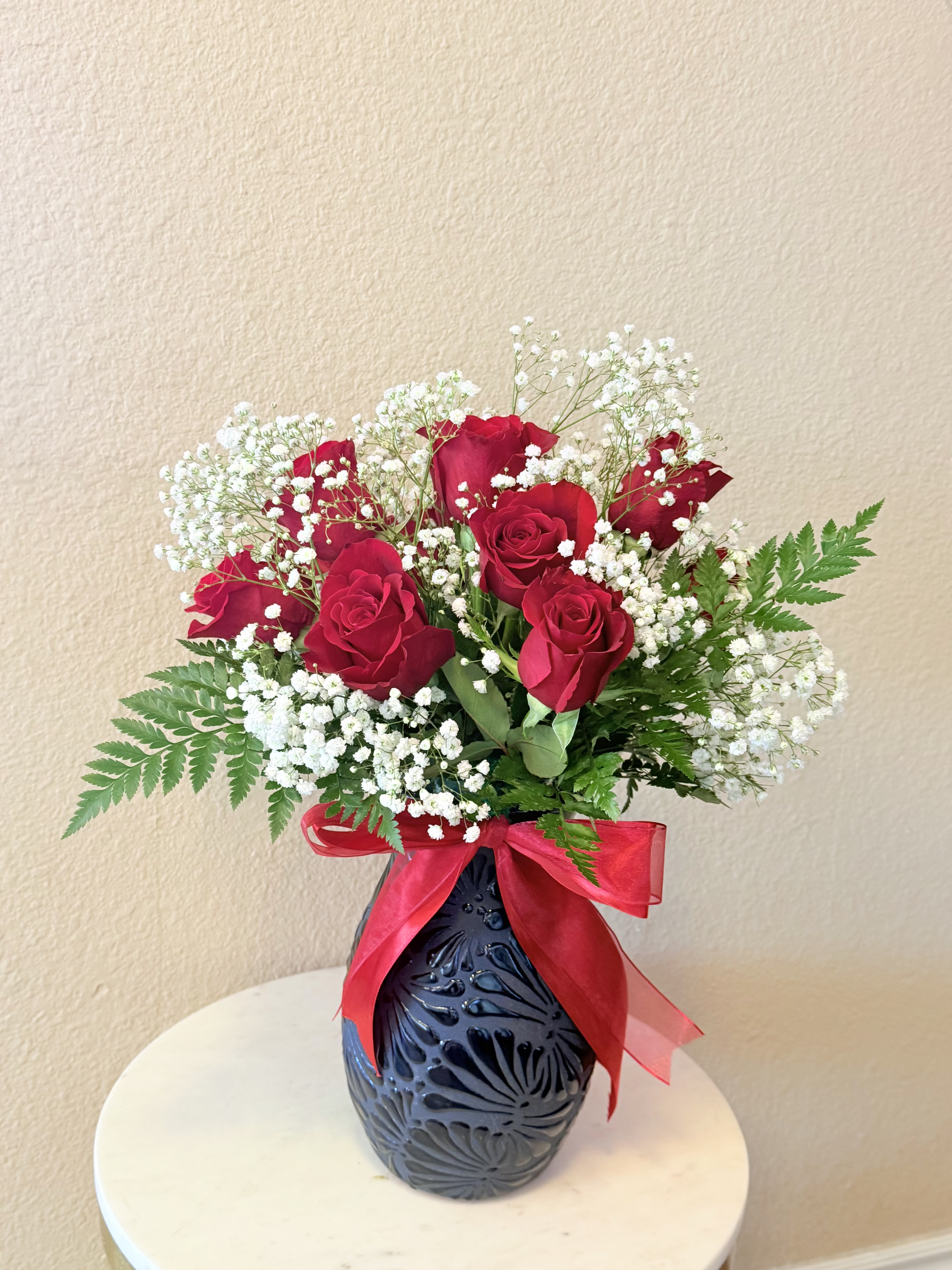 Endless Love for Mom - Deep, velvety red roses clustered with clouds of white baby's breath create a timeless, romantic statement designed to make Mom feel cherished on Mother's Day, an anniversary, or just because. The arrangement reads classic and passionate at first glance-the saturated red blooms send a clear message of love while the delicate white filler softens the mood so she feels both celebrated and comforted.  Set in a dark, textured ceramic vase tied with a bright red ribbon (and accented by an additional pink ribbon detail), this full, rounded bouquet uses fern-like greenery to add airy movement and a soft, natural frame. The contrast of red, white, green and the black-toned vase gives the display strong visual impact on mantels, dining tables, or bedside stands. Sized to be clearly seen but still intimate, the piece highlights roses as the focal flower with baby's breath providing a cloudlike backdrop and supportive texture.  Perfect for the mom who delights in classic romance, this arrangement is the kind of gift that reads as both grand and personal-the sort of bouquet that makes her feel truly seen and loved. Easy care: keep water fresh and trim stems every few days to extend vase life. Order with confidence for same-week delivery when you need a heartfelt surprise.