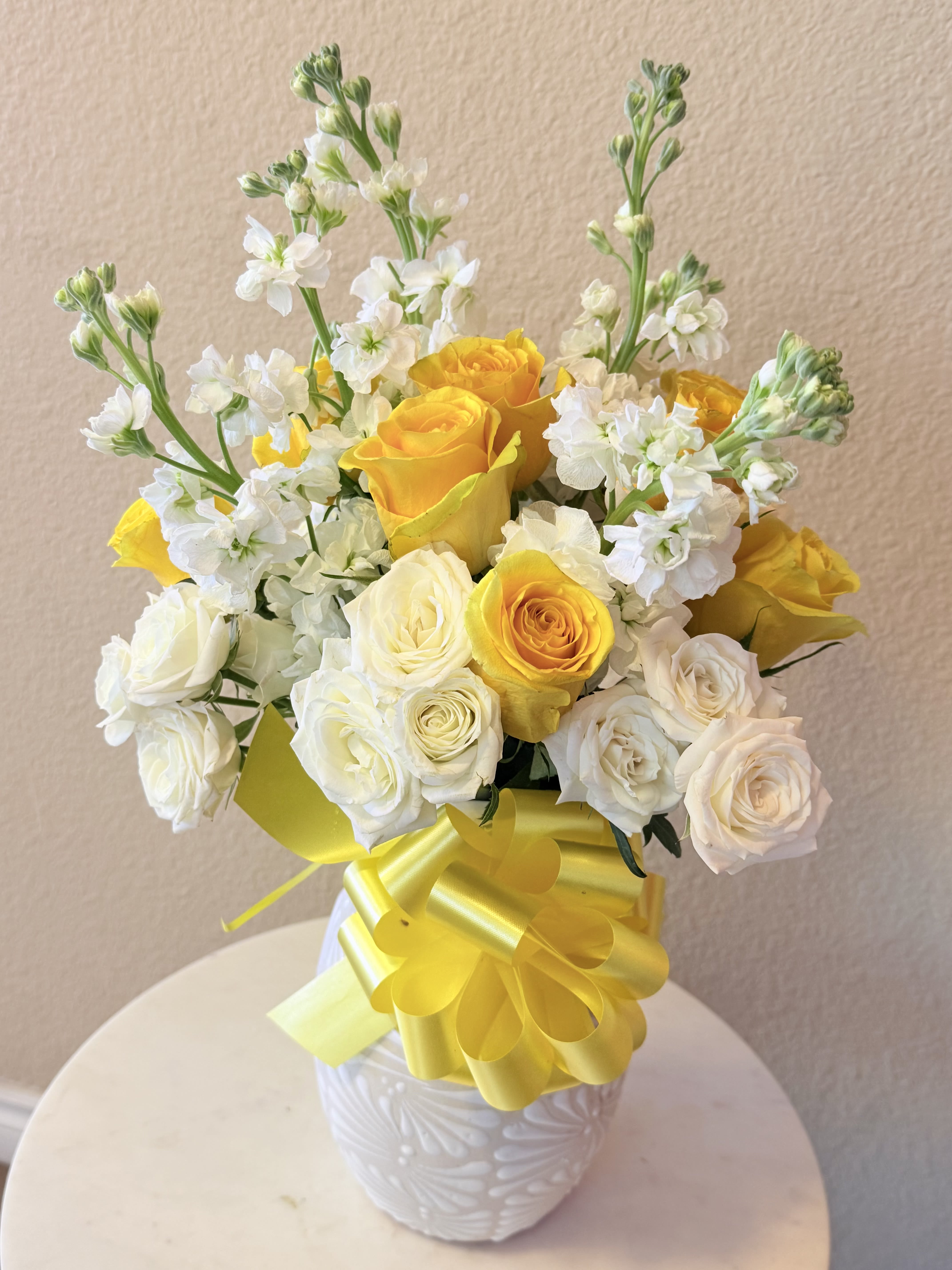 Sunshine for Mom - Sunlit yellow roses mingle with soft white roses and tall white stock to create a cheerful birthday bouquet or spring celebration arrangement that feels bright, fresh, and hopeful. The yellow-and-white color story and the upward sweep of the stock give the recipient an immediate sense of warmth and uplift-the kind of bouquet that makes someone feel truly celebrated or specially thought of.  Dense, rounded clusters of blooms sit in a white ceramic vase tied with a broad yellow ribbon for a polished, gift-ready presentation. The composition balances compact, velvety rose heads with the airy texture and height of stock, delivering both visual impact and a gentle garden-style feel. Ideal for birthdays, thank-you moments, or just-because springtime surprises; vase-ready and arranged for lasting freshness-refresh water regularly and recut stems if transferring to a different container.