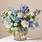 Blue Sky And Sunshine  - Soft blue delphinium spikes rising above a cloud of white roses give this floral arrangement a serene, uplifting presence-perfect as a Mother's Day bouquet or a thoughtful birthday surprise. The flower arrangement pairs pale blue hydrangea-like blooms and airy lisianthus with layered white roses, creating a calm, elegant palette that feels both fresh and refined for everyday gifting.  Arranged in a clear glass vase and finished with a light ribbon, the composition is full and rounded yet airy, with silvery-green foliage adding subtle contrast and texture. Expect a lush, polished look that reads as relaxed sophistication; it's the kind of bouquet that makes the person you're thinking of feel truly seen and cared for without being over the top. Standard vase dimensions and simple care-fresh water and a cool spot away from direct sun-will help the blooms stay beautiful through the week.