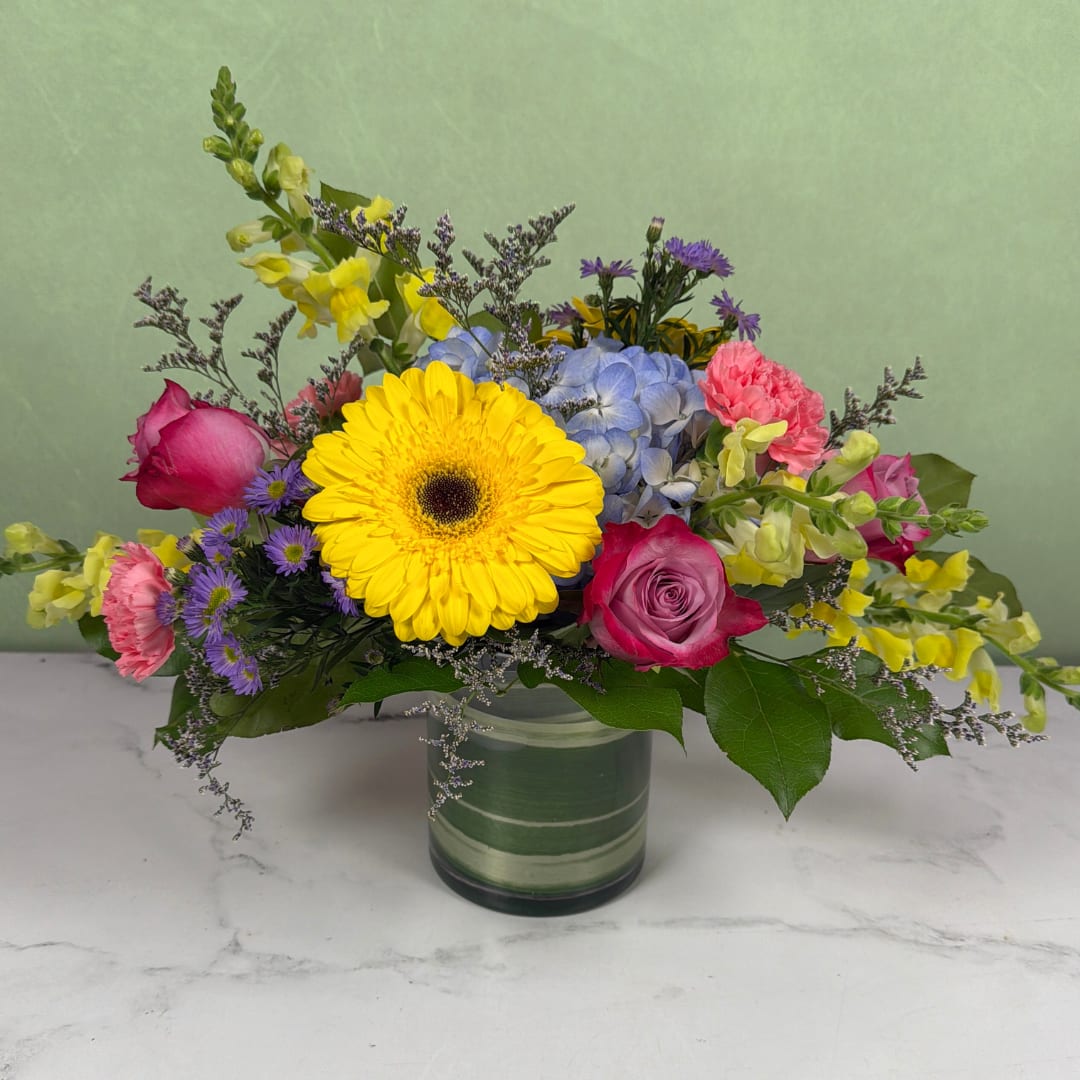 Breath of Spring Cylinder - Colorful flowers are blooming in this Breath of Spring Cylinder by Savilles Florist, making it an instantly cheerful choice for a birthday bouquet, get well flowers, or a thinking-of-you gift. Deep purple roses and pink carnations surround yellow gerbera daisies, balanced by soft blue hydrangea, sunny yellow snapdragons, and tiny purple aster accents for contrast and texture.   The stems are placed in a clear cylindrical vase. and the low, wide design creates a garden-fresh, playful feel that will make the recipient smile the moment it arrives. Compact but generous in presence, this arrangement is perfect for a table centerpiece or brightening a bedside or desk.  Ideal for sending congratulations, thanks, or encouragement, this arrangement is made to feel casual and celebratory, and is the kind of bouquet that makes someone feel genuinely seen. Arrives ready to display in the included glass cylinder; refresh water every few days and trim stems if desired to extend freshness.