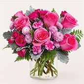 Sweet as Can Be - A lush cluster of bright pink roses and soft pink carnations creates an instantly cheerful, romantic statement - perfect as a birthday bouquet, anniversary arrangement, or Mother's Day gift to let someone know you're thinking of them. The rounded, densely packed shape reads feminine and full, so the recipient will feel truly celebrated the moment they see it.  This flower arrangement pairs varying shades of pink roses with smaller pink carnations and silvery foliage for contrast, all gathered into a classic clear glass vase for an evergreen, giftable presentation. The compact, rounded design emphasizes abundant blooms and a soft texture; green stems visible through the vase add a fresh, natural touch that complements the pink-and-white palette.  Ideal for everyday gifting when you want to brighten someone's day, send thanks, or mark a special milestone, this arrangement arrives ready to display on a table, desk, or bedside. Ask about size upgrades at checkout for a larger display; keep the water fresh and trim stems every few days to help the roses and carnations last longer.