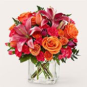 Lucky Lily - A dazzling cluster of pink lilies and vibrant orange roses makes a cheerful, romantic statement-perfect as a birthday bouquet, anniversary arrangement, or a bright thank-you gift. The lively mix of reds, corals, and whites woven through the composition gives the person you're thinking of an energetic, affectionate impression the moment the vase is set on their table.  Dense, rounded and full, the Lucky Lily arrangement pairs fragrant pink lilies with saturated orange roses and bright red carnations, all closely packed for a lush, layered texture. Smaller coral and white blooms tuck into the mix for contrast, and the clear square glass vase keeps the look clean and modern while showcasing the stems and structure.  This is the kind of everyday gift that reads as both celebratory and sincerely thoughtful-send it to mark a milestone or simply to make someone feel truly seen. Arrives ready to display in a clear glass vase; for best enjoyment, change the water every 2-3 days and trim stems slightly to extend the life of the blooms.