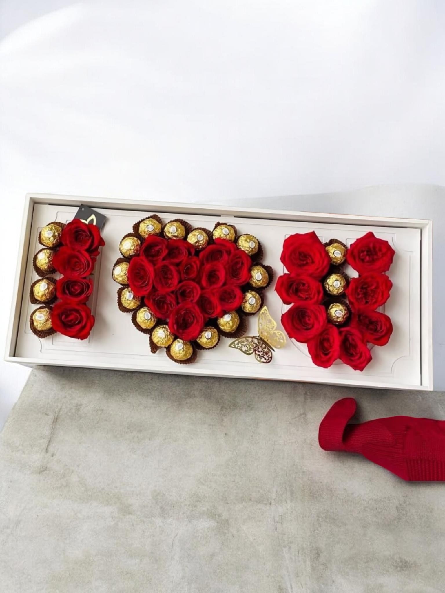 Love Box - Deep red roses formed into heart-shaped clusters make an immediate romantic statement-perfect as a Valentine's Day bouquet or anniversary arrangement that says "I love you" without words. The long rectangular gift box presentation gives this floral gift a polished, keepsake feel, and the finished look is designed to make the recipient feel truly cherished.  Each arrangement pairs velvety red roses with gold-wrapped Ferrero Rocher chocolates tucked throughout the box, the metallic highlights playing beautifully against the rich petals and dark bases. A decorative butterfly embellishment and ribbon add a delicate, celebratory touch, while the compact rectangular size makes it ideal for delivery or handoff in person.  Choose this for the person you want to sweep off their feet-it's the kind of gift that reads as both intimate and festive for anniversaries, Valentine's Day, or any romantic milestone. Arrangement dimensions and care instructions are provided at checkout to help you select the best size and ensure the roses stay fresh.