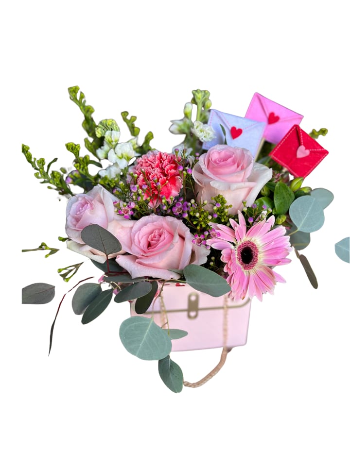 Beverly Hills - Soft pale pink roses and a single pink gerbera daisy create the romantic center of this flower arrangement, perfect as a birthday bouquet, anniversary roses alternative, or a cheerful Valentine's Day gift. The compact, gift-like design feels playful and affectionate-she'll feel genuinely celebrated when those heart-detailed envelope picks peek out from the back, adding a sweet, personal touch.  A cluster of bright pink carnations and small purple accent blooms surround the focal roses, balanced by airy eucalyptus-like greenery for texture and movement. Arranged in a pink container, the mix of pinks, whites and touches of purple reads soft and feminine without being fussy; proportions are deliberately compact for easy display on a bedside table or desk.  This arrangement is ideal for saying "I'm thinking of you" to a partner, friend, or family member who loves a romantic, whimsical look. It's delivered ready to display in its decorative container-minimal care needed beyond fresh water if you transfer stems-making it an effortless, heartfelt surprise.