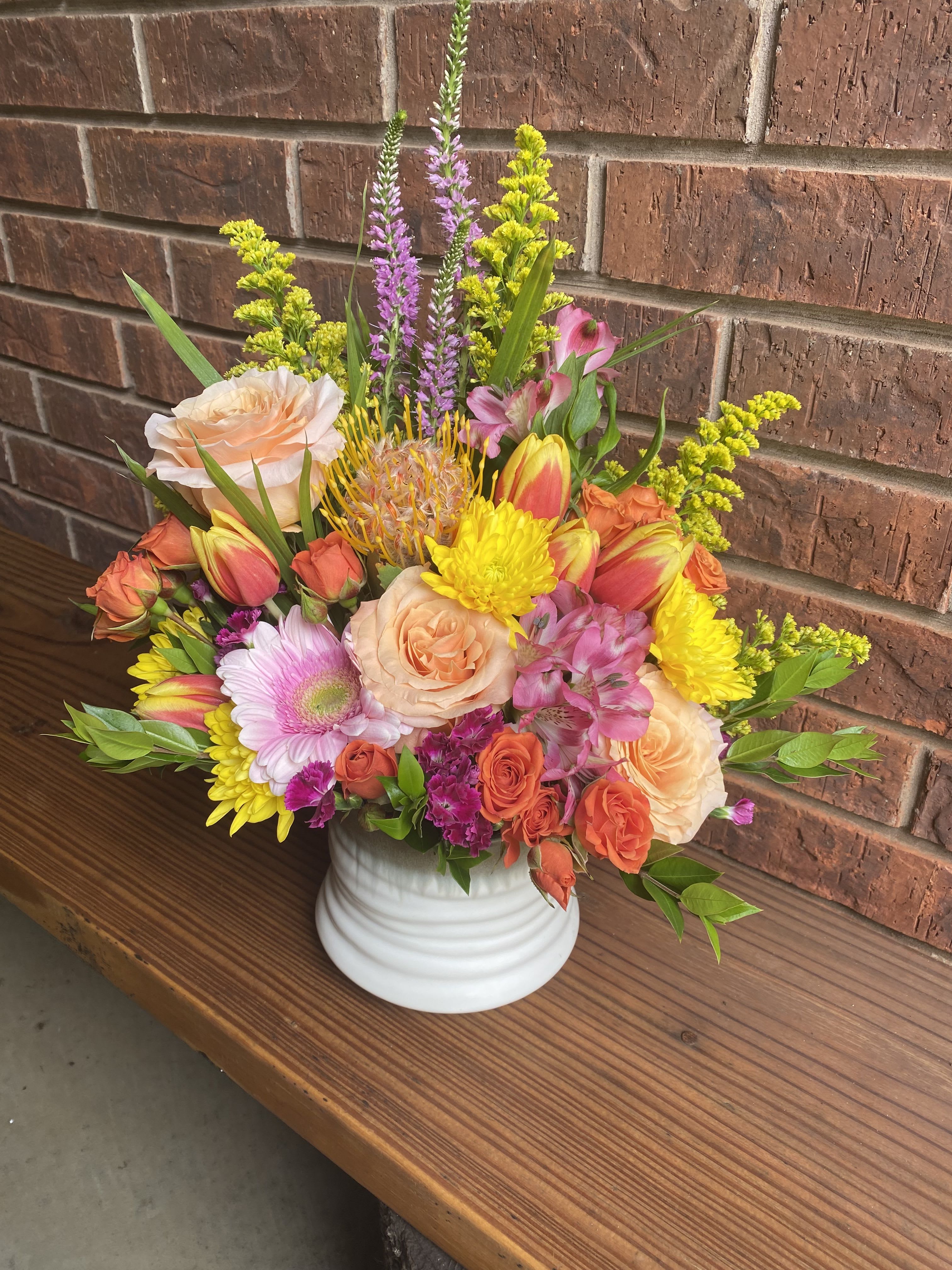Cheer-io - A burst of sunlit color and modern texture-this cheerful flower arrangement is a birthday bouquet and celebration centerpiece all in one. The rounded composition of peach and orange roses with sunny yellow chrysanthemums reads joyful and uplifting, while pink gerbera daisies and alstroemeria bring a playful, feminine touch that makes the recipient feel truly celebrated.  The mix includes peach roses, orange spray roses, yellow chrysanthemums, pink gerbera daisies, alstroemeria, bicolor tulips, and a spiky pincushion protea that creates a contemporary focal point. Vertical purple flower spikes and small yellow filler blooms add height and movement above the dense central cluster. Everything is arranged in a simple white ceramic vase for clean contrast and easy display.  Perfect for birthdays, congratulations, or "just because" moments, this arrangement is made to make someone feel bright and noticed-ideal for coworkers, friends, or family. Vase-ready on delivery; refresh with a few inches of water and recut stems every few days to extend the life of the flowers.