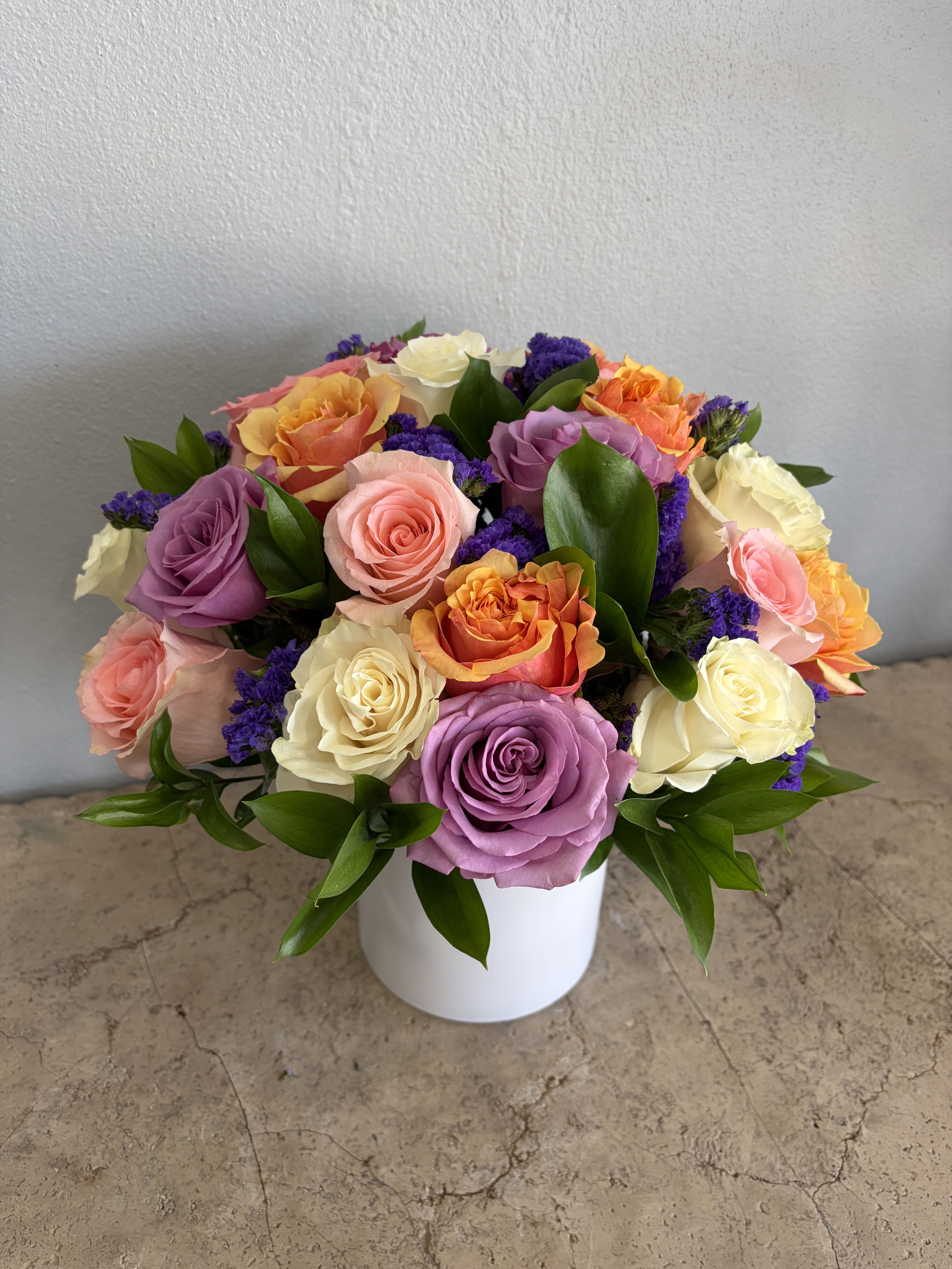 Carnival - The CARNIVAL bouquet features a beautiful variety Roses in shades of peach, orange, white and purple. Accented with green Ruscus and Purple statice designed in a white ceramic vase. Available for pickup or delivery in West Palm Beach, Palm Beach, Royal Palm Beach, Palm Beach Gardens and surrounding areas.