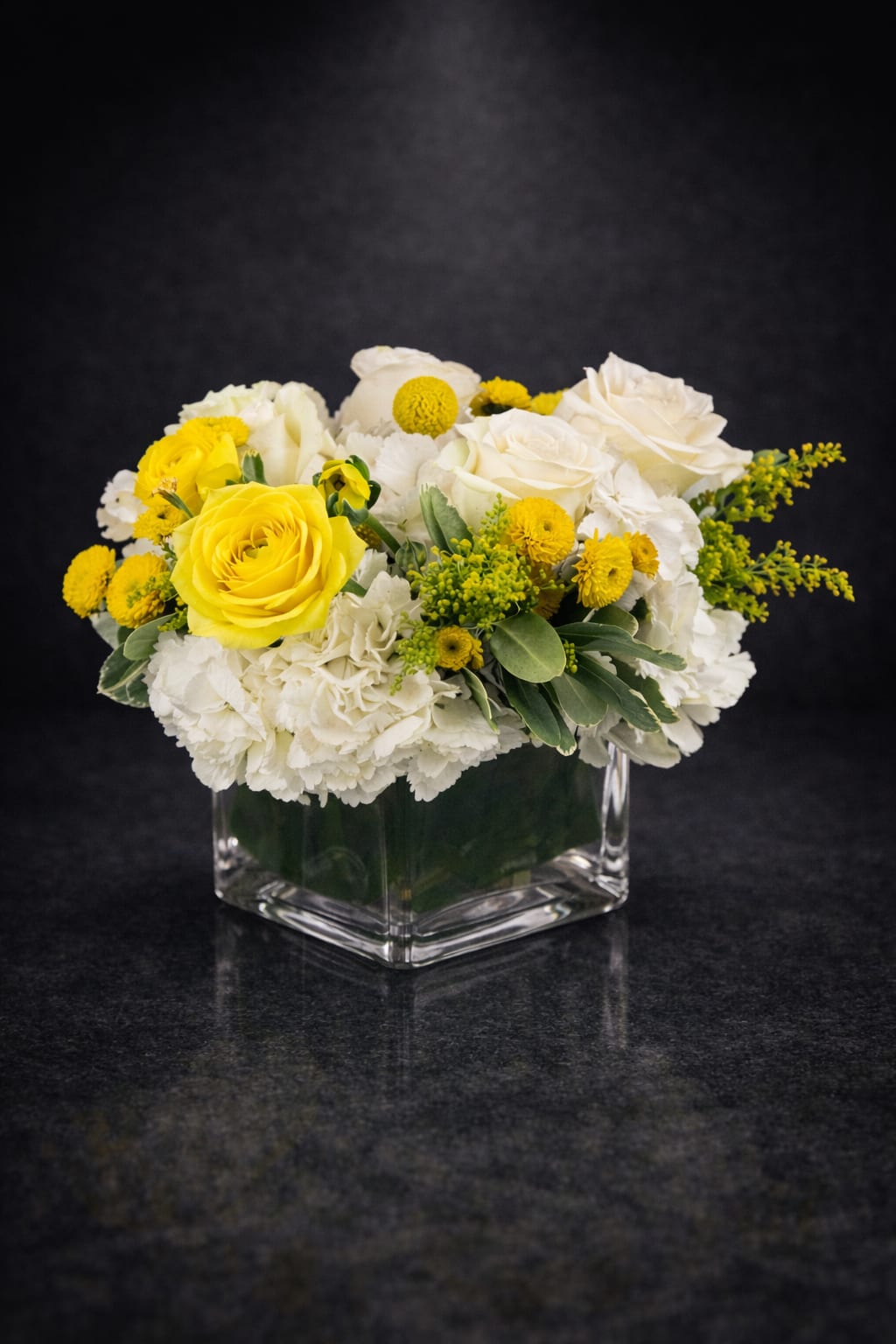 Golden Ivory - Sunlit yellow roses paired with creamy white roses and clustered white carnations create an uplifting everyday flower arrangement perfect for a birthday bouquet, a thank-you gift, or a fresh spring celebration. The bright yellow blooms instantly brighten the room while the soft white base and green filler give the recipient a feeling of warmth and calm-she'll feel genuinely celebrated and noticed when these arrive.  This low, compact design sits in a clear square glass vase for a modern, clean presentation that fits on coffee tables, desks, or kitchen counters. Small yellow button-like blooms and sprigs of green filler add playful texture and contrast against the mostly white palette. The arrangement's cheerful color mix and tidy proportions make it an easy, elegant choice for sending bright wishes or saying "I'm thinking of you" with style.