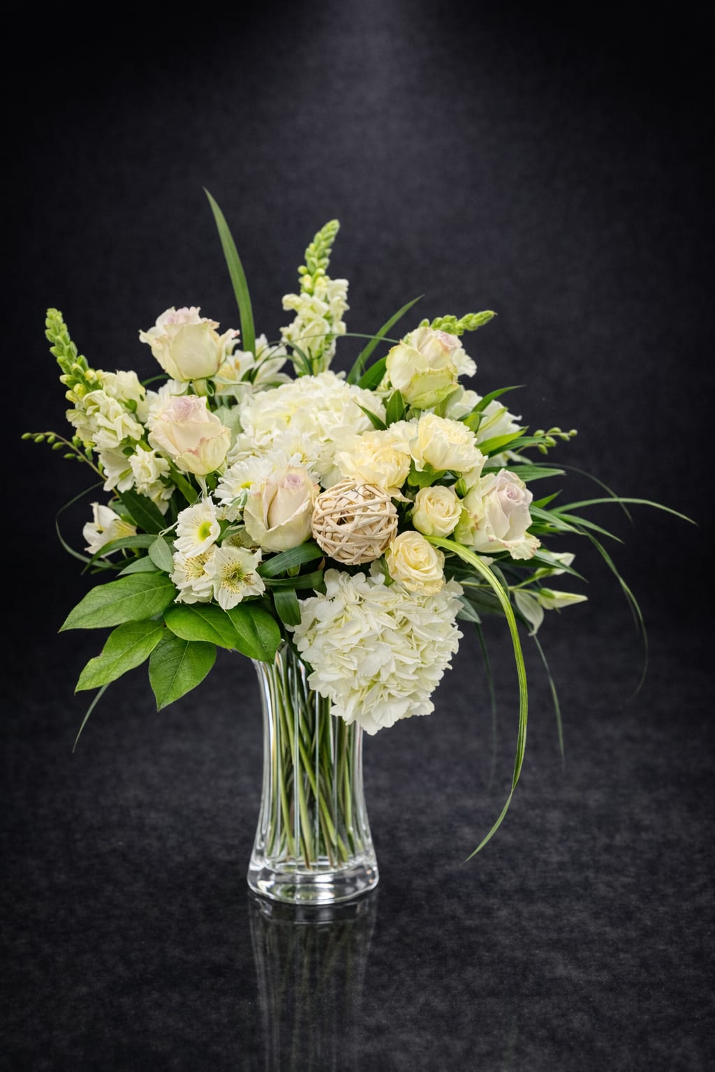 White Whisper - Soft white roses and pale blush hydrangeas give this flower arrangement a serene, garden-fresh presence-perfect as a sympathy bouquet, a thoughtful thank-you gift, or a springtime surprise for someone who loves gentle colors. The arrangement's airy silhouette and long, arching greenery make the recipient feel quietly comforted and seen, the kind of bouquet that reads as both elegant and effortlessly comforting.  Clustered roses, fluffy hydrangeas, tall snapdragons, delicate alstroemeria, and cheerful daisies are grouped in a clear glass vase, accented by broad leaves and a decorative wicker ball tucked near the center for a natural, textural touch. Colors are white, soft green, and blush pink; the overall shape is loose and organic to suggest freshly gathered garden blooms. Designed for everyday gifting as well as more tender moments, it arrives ready to display-keep stems trimmed and the water fresh to extend the life of the flowers.