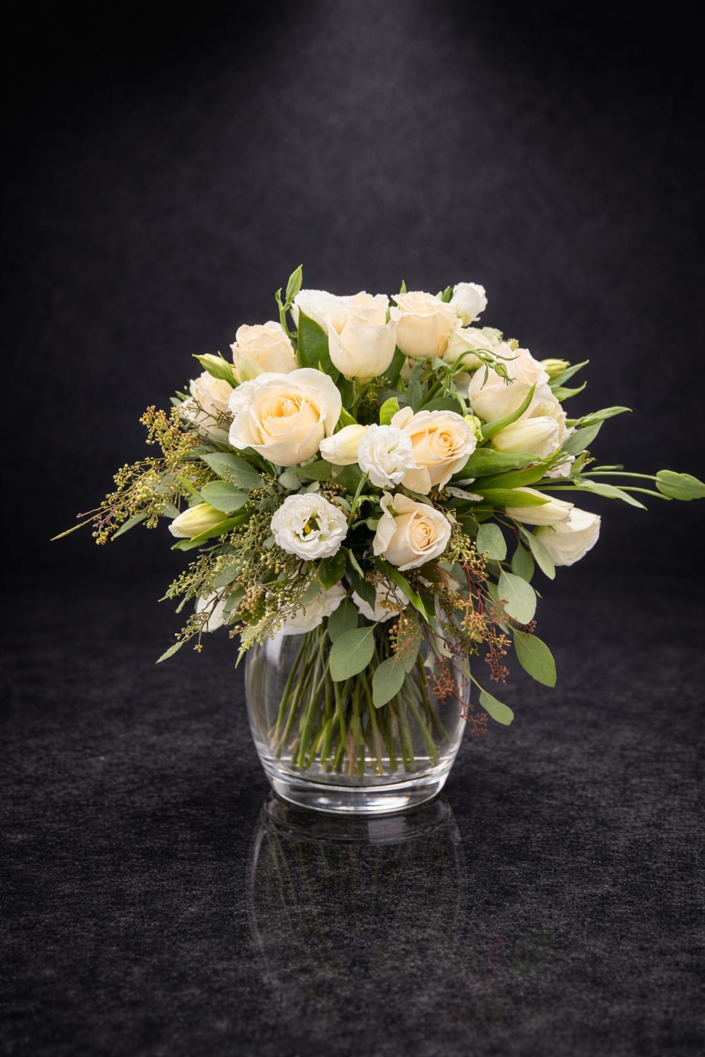 Cream Elegance - Cream-toned roses and white lisianthus mingle with airy greenery in a rounded bouquet that reads soft, romantic, and quietly refined-perfect as an anniversary bouquet, a sympathy arrangement, or a thoughtful thank-you gift. The gentle ivory and blush-peach petals create a calming presence that will make the person you're thinking of feel seen and comforted without fuss.  Arranged in a clear glass vase, the composition keeps the focus on texture and color: full roses provide plush, velvety blooms while the lisianthus adds delicate, ruffled contrast and lightness. The palette of white, peach, and green suits both classic and contemporary settings; the vase's clean lines make it easy to place on a mantle, bedside table, or entry console.  Choose this arrangement when you want to convey warmth and understated elegance-whether marking an anniversary, offering condolences, or saying thanks. Standard dimensions fit most tabletops; to keep blooms fresh, change the water every two days and trim stems slightly when you refresh the vase.