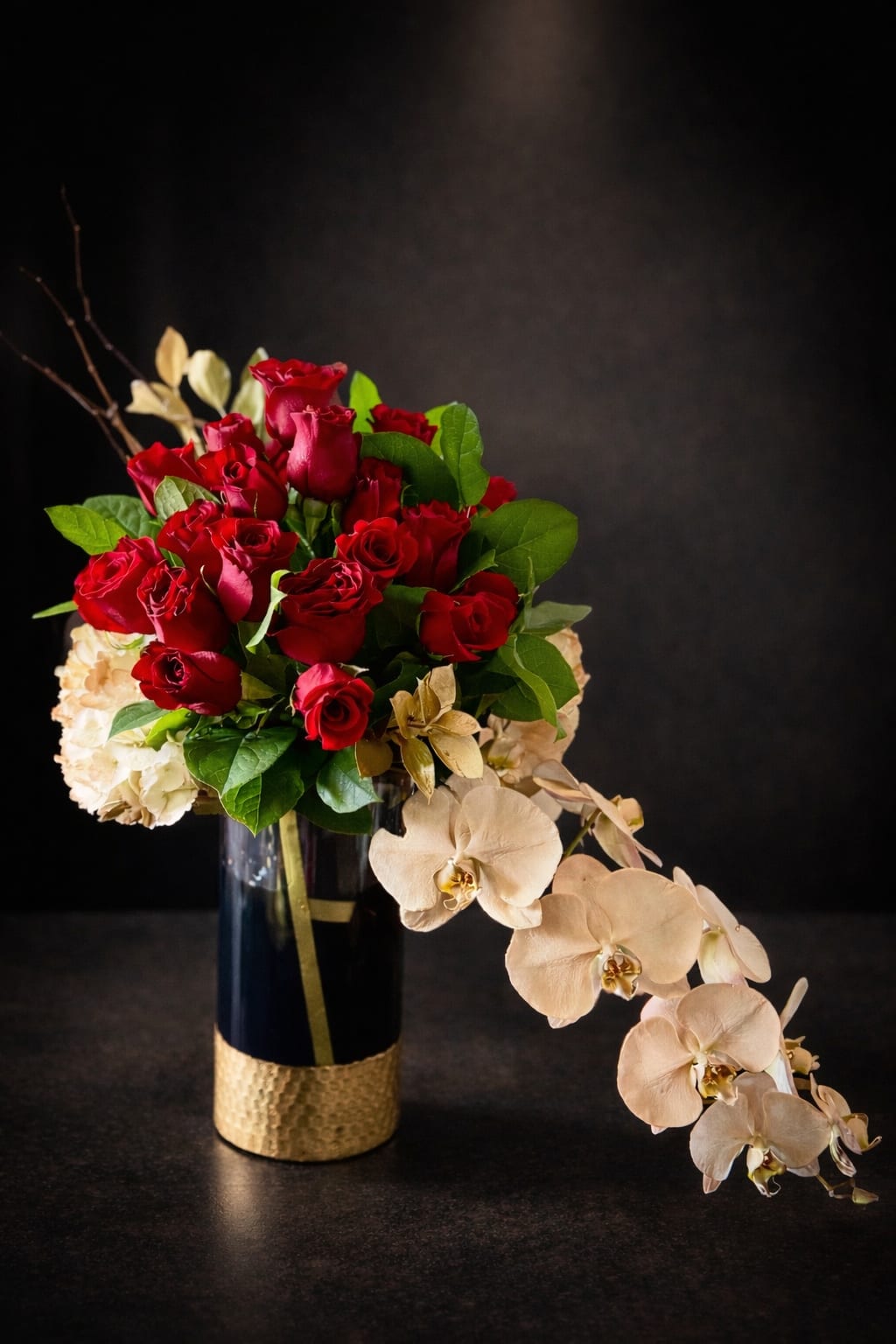 Velvet Orchid - A dramatic cluster of velvety red roses crowns this floral arrangement, accented by creamy white hydrangeas and a trailing stem of pale phalaenopsis orchids-the ideal anniversary bouquet or romantic gift that reads both elegant and intimate. As a flower arrangement for love and romance or a thoughtful thank-you, Velvet Orchid makes the person you're thinking of feel deeply seen: the red-and-ivory contrast is strikingly romantic, while the delicate orchid cascade lends a gentle, graceful movement.  Set in a tall, dark glass vase with a textured gold base and finished with a narrow gold ribbon, this design feels polished and formal without being fussy. The composition combines rich red roses, soft cream hydrangea clusters, and pale Phalaenopsis orchids in a compact, sculpted silhouette with a cascading accent; colors include red, white, green foliage, and a touch of black from the vase's deep tone. The vase height and structured shape give it presence on a dining table or entry console, making it a refined everyday arrangement that works for anniversaries, romantic surprises, or to express sincere thanks.