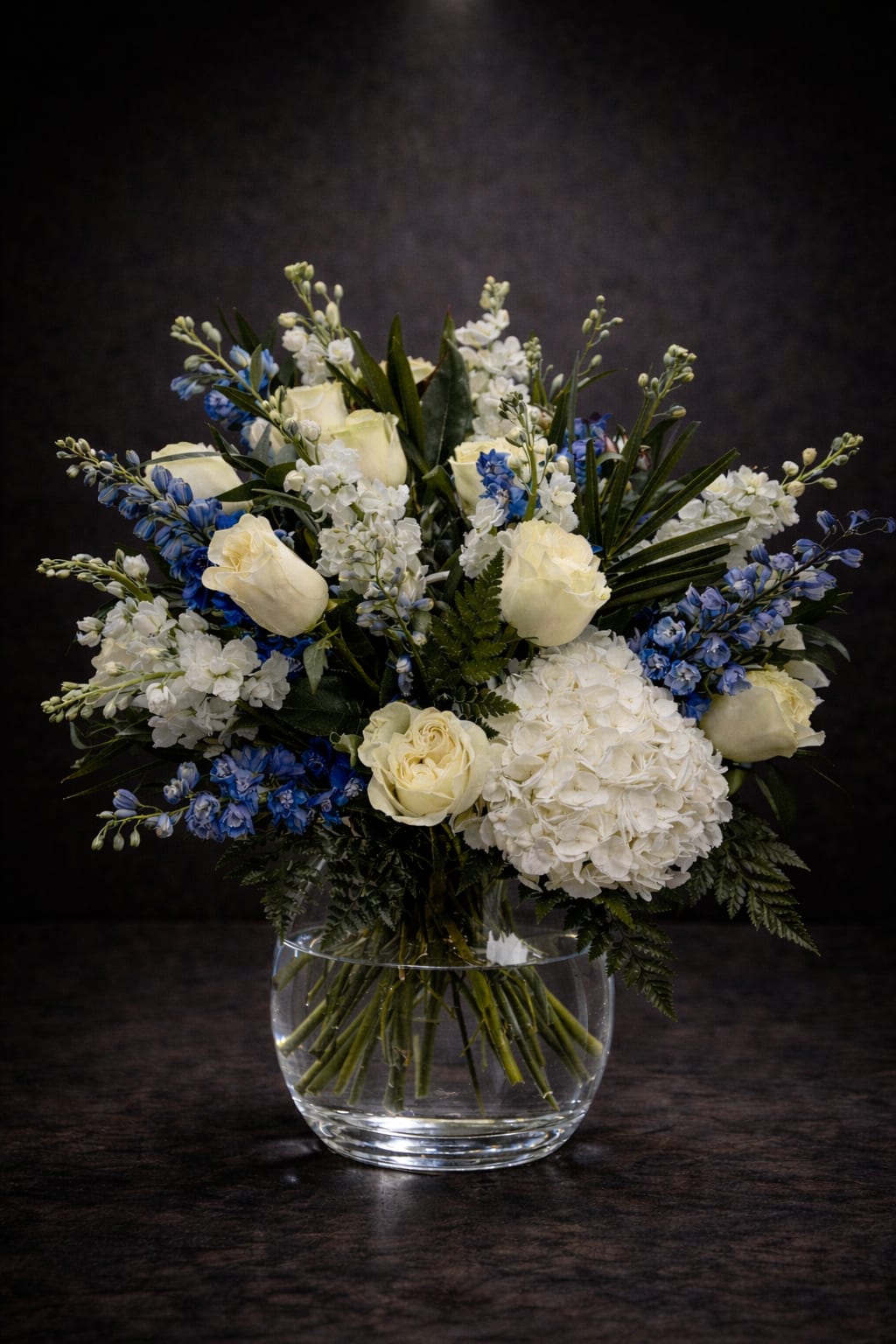 Sky Elegance - Creamy white roses and lush white hydrangeas meet vivid blue florals in a cool, elegant flower arrangement perfect as a sympathy arrangement, a thoughtful thank-you, or a serene everyday gift. Sky Elegance is designed to make the person you're thinking of feel calm and cared for-the soft whites read as gentle and sincere, while the blue clusters add a refined, peaceful note that quietly uplifts.  This full, rounded arrangement layers textures-velvety rose petals, billowy hydrangea heads, and upright blue accents-for a balanced, formal silhouette. Arranged in a clear glass vase that reveals the stemwork at the base, the presentation stays simple and sophisticated, allowing the flowers' shapes and colors to take center stage. The color palette of white, blue, and green keeps the look timeless and versatile for many occasions.  Ideal for offering comfort, expressing gratitude, or marking a quiet celebration, Sky Elegance arrives ready to display on a mantel, bedside table, or reception desk. Care is straightforward: fresh water in the vase and a fresh cut to the stems every few days will help maintain the arrangement's serene, just-arranged appearance.