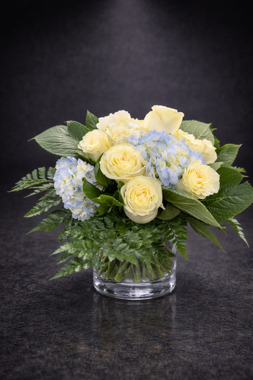Ivory Bloom - Pale cream roses paired with soft blue hydrangeas create a calm, elegant centerpiece that reads as fresh and understated - perfect as a birthday bouquet, a thank-you arrangement, a spring celebration accent, or a sympathy display. The rounded, compact shape feels soft and balanced, so the person you're sending it to will feel seen and comforted by its gentle, reassuring presence.  Cream roses and light blue hydrangea clusters sit low in a clear glass vase, framed by broad green foliage that adds contrast and a touch of natural texture. The cool pastel palette and modest scale make it versatile for a mantel, bedside table, or dining centerpiece; arranged densely for a lush look, it keeps a neat, contemporary silhouette suitable for everyday gifting or more solemn moments. Care: keep water fresh and trim stems every few days to prolong bloom life.