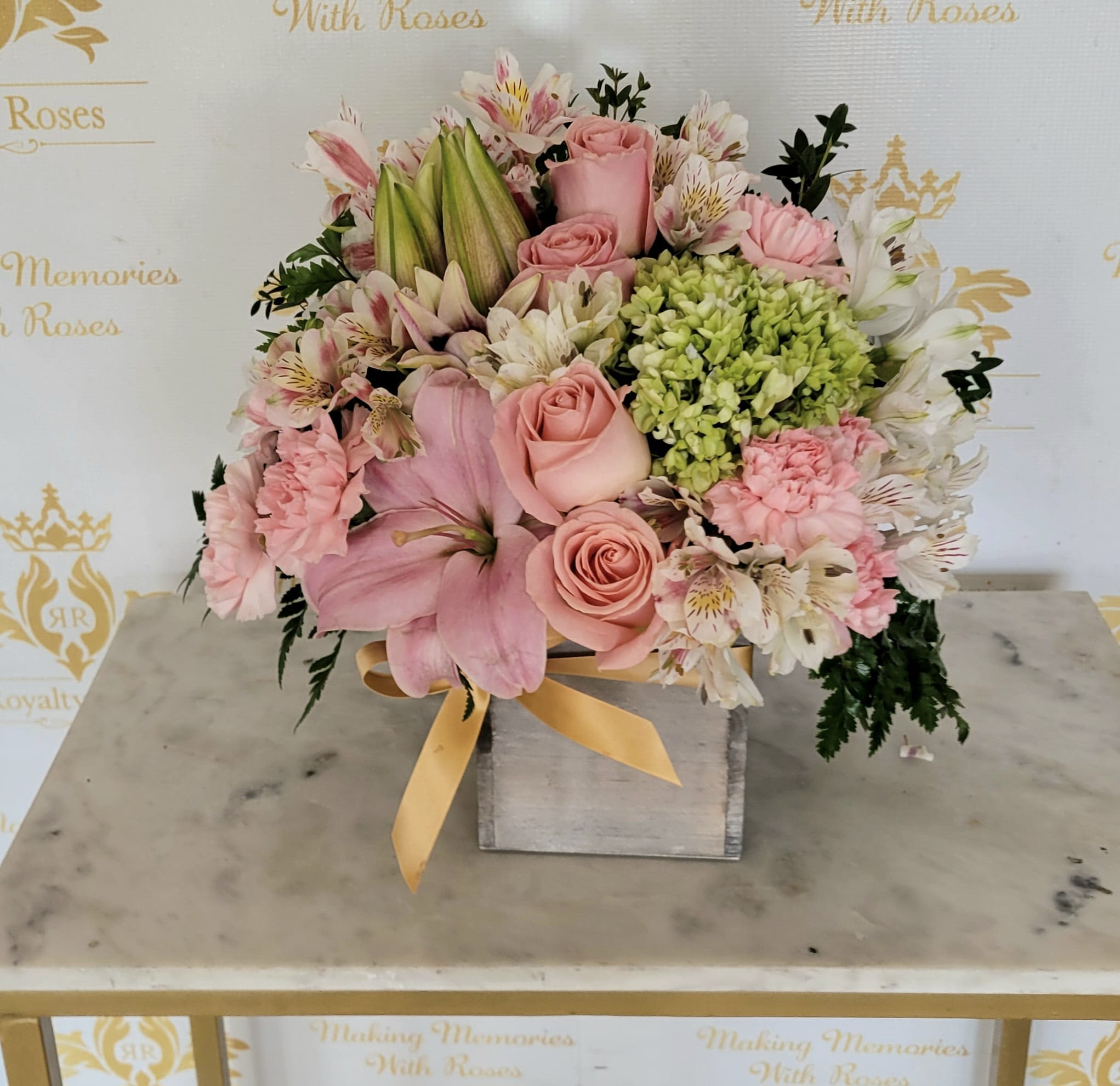 All about pink - Soft pink roses and lilies take center stage in this All about pink flower arrangement, a gentle birthday or anniversary bouquet that also reads thoughtful for Mother's Day or sympathy. The mix of pale pink carnations and white alstroemeria brings delicate texture while the light green hydrangea adds a fresh, airy contrast-she'll feel comforted and celebrated the moment she sees it.  The flowers are artfully arranged and snugly packed into a square wooden box, finished with a yellow satin ribbon for a polished, gift-ready presentation. Colors include soft pink, white, green, and a touch of sunny yellow from the ribbon; blooms include roses, lilies, alstroemeria, carnations, and hydrangeas for a layered, romantic look with varied shapes and scent.  Perfect for the person you want to make feel truly seen-whether marking a milestone, offering sympathy, or saying "thank you." The compact wooden box makes it easy to display on a table or desk; order with standard delivery to ensure peak freshness on the day you choose.