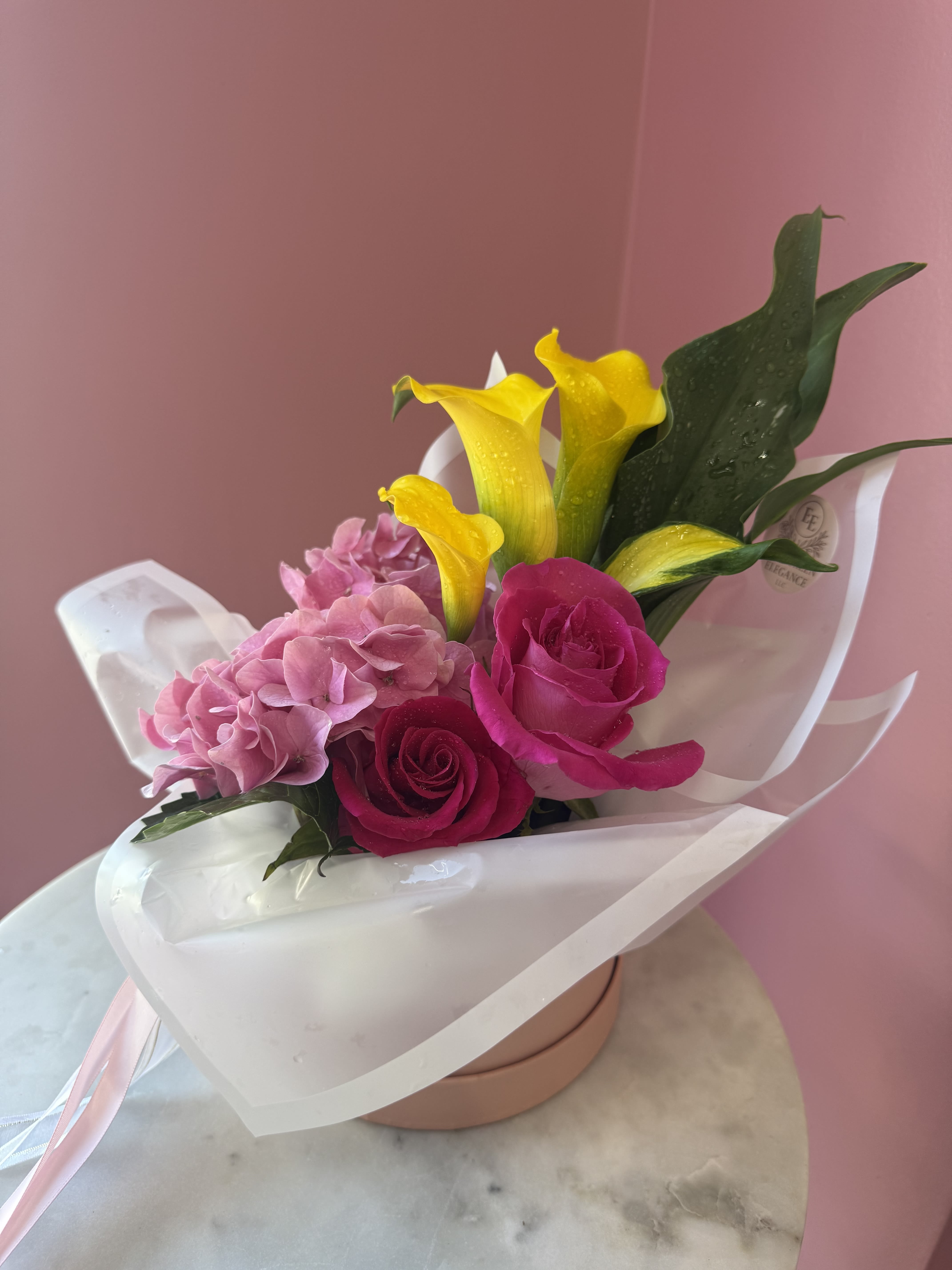 Calla Lilly Spring  - Bright pink roses and vivid yellow calla lilies stake a joyful claim in this spring bouquet, making it a perfect birthday bouquet or romantic gift that reads as cheerful and celebratory. The combination of soft pink hydrangea with the punch of yellow creates a lively contrast that will make the person you're thinking of feel delighted and seen-ideal for congratulations, anniversaries, or simply to brighten someone's day.  This compact hand-tied arrangement is wrapped in glossy white paper and finished with a pale pink ribbon, presented in a round pink box for an instant gift-ready moment. Expect a lush mix of hydrangeas, roses, and calla lilies in pink, yellow, and white tones; the close, rounded shape emphasizes texture and color contrast while staying easy to carry and display. Simple care-fresh water in the box or a vase and a cool spot out of direct sun-keeps the blooms looking their best for several days.